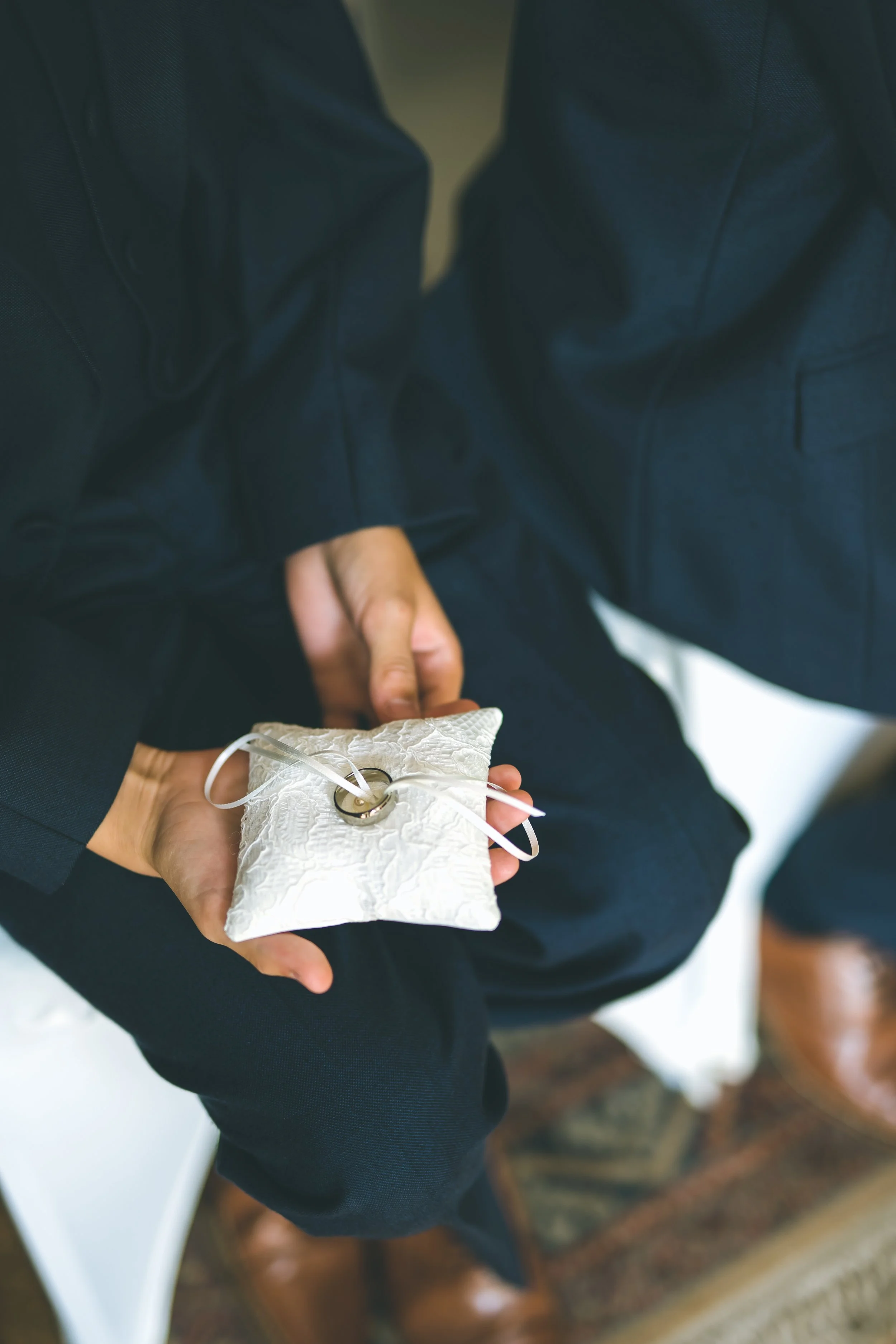 Person in formal attire holding a wedding ring on a white decorative pillow during a marriage ceremony.