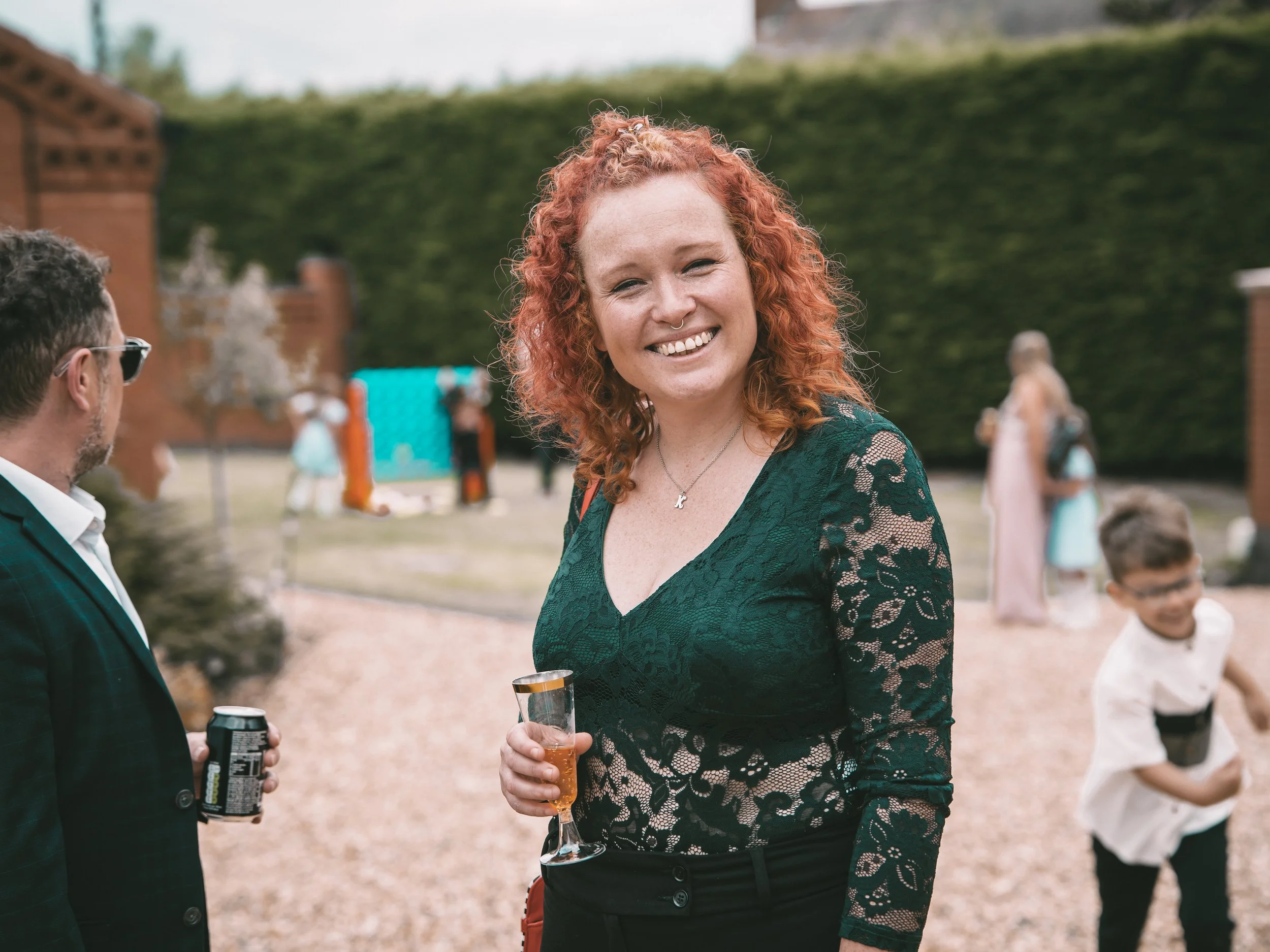 A woman with curly red hair smiling outdoors during daytime, holding a glass of wine in one hand and a beverage in the other, wearing a dark green lace top.