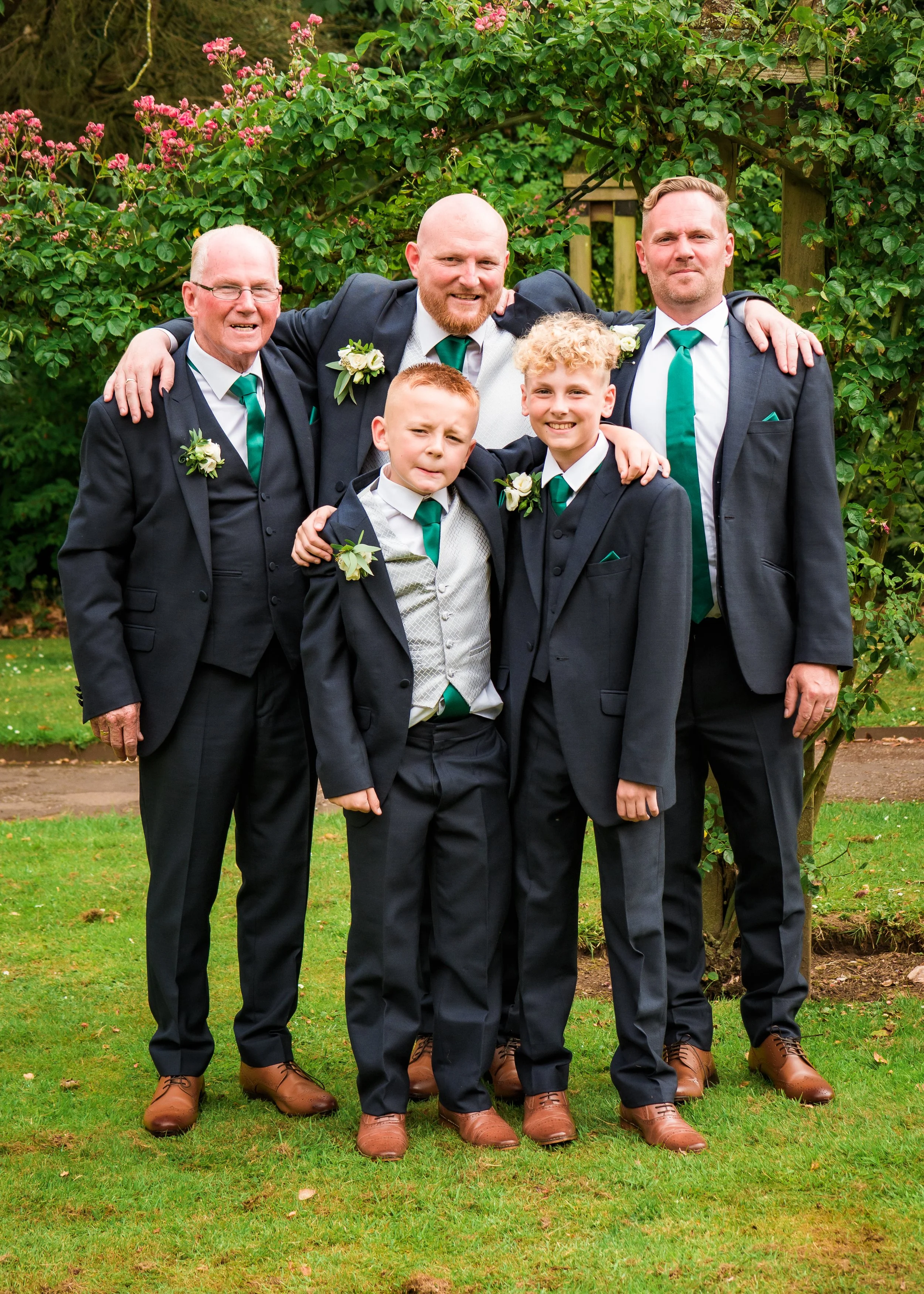 Five men and two boys in formal suits posing outside in a garden with green shrubs and pink flowers, all wearing dark suits with white shirts, green ties, and boutonnières, standing close together with arms around each other.