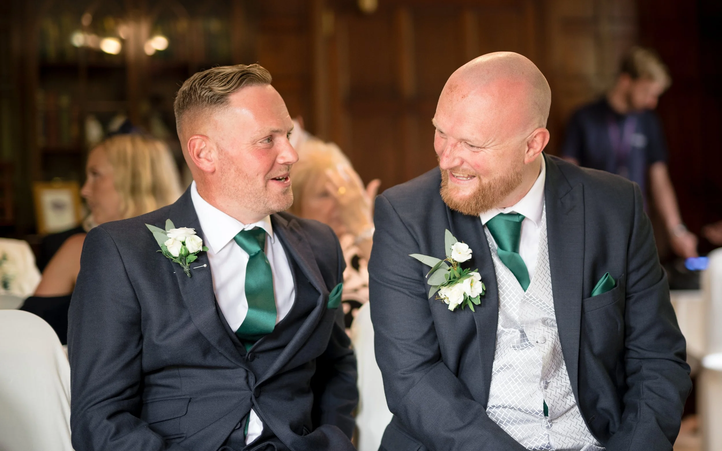 Two men in formal suits and green ties sitting close together, smiling and engaging in conversation at a wedding or formal event. Both are wearing white floral boutonnieres.