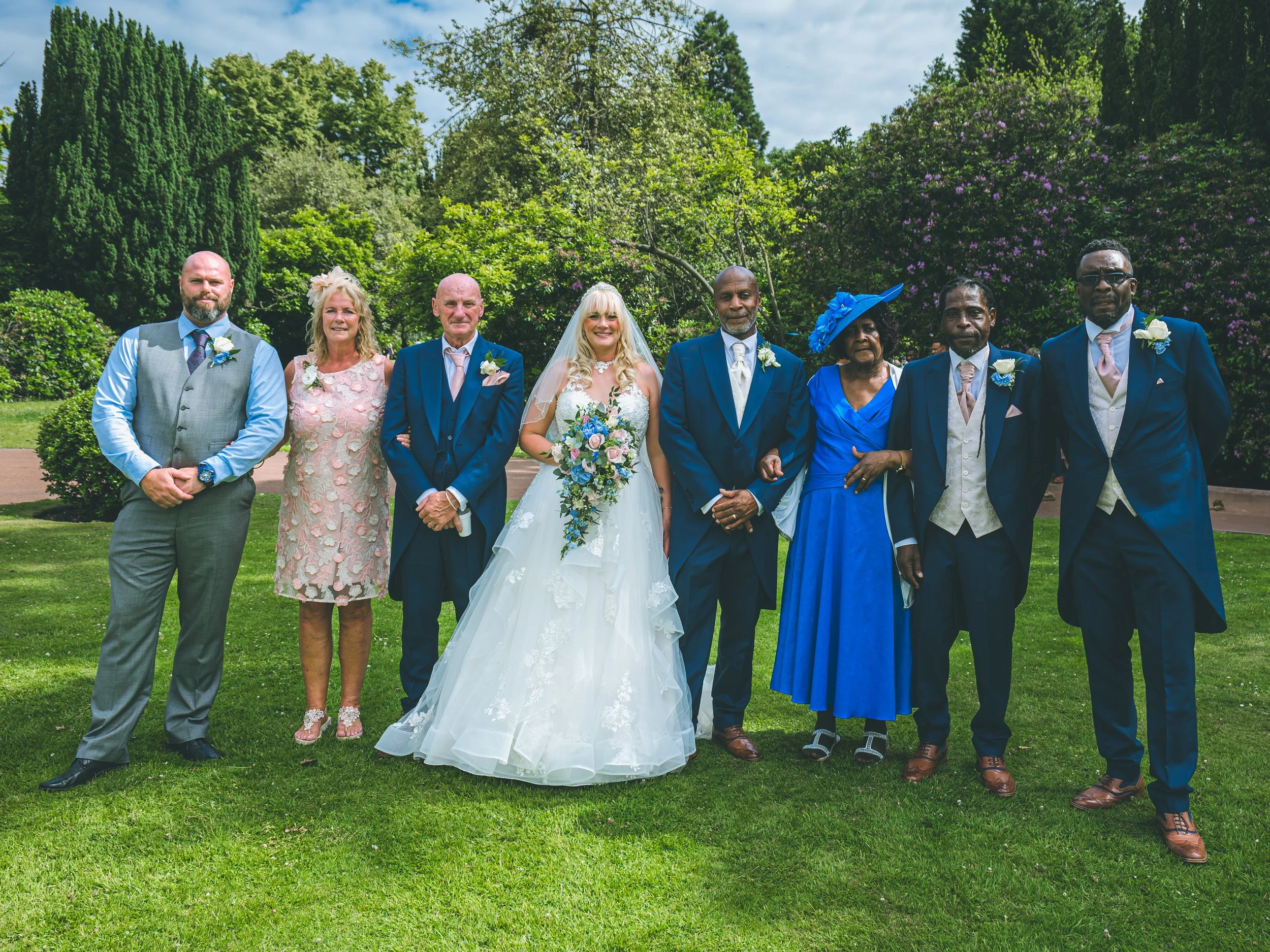 Group of seven diverse people standing outdoors on grass, dressed in formal attire, attending a wedding, with trees and bushes in the background.