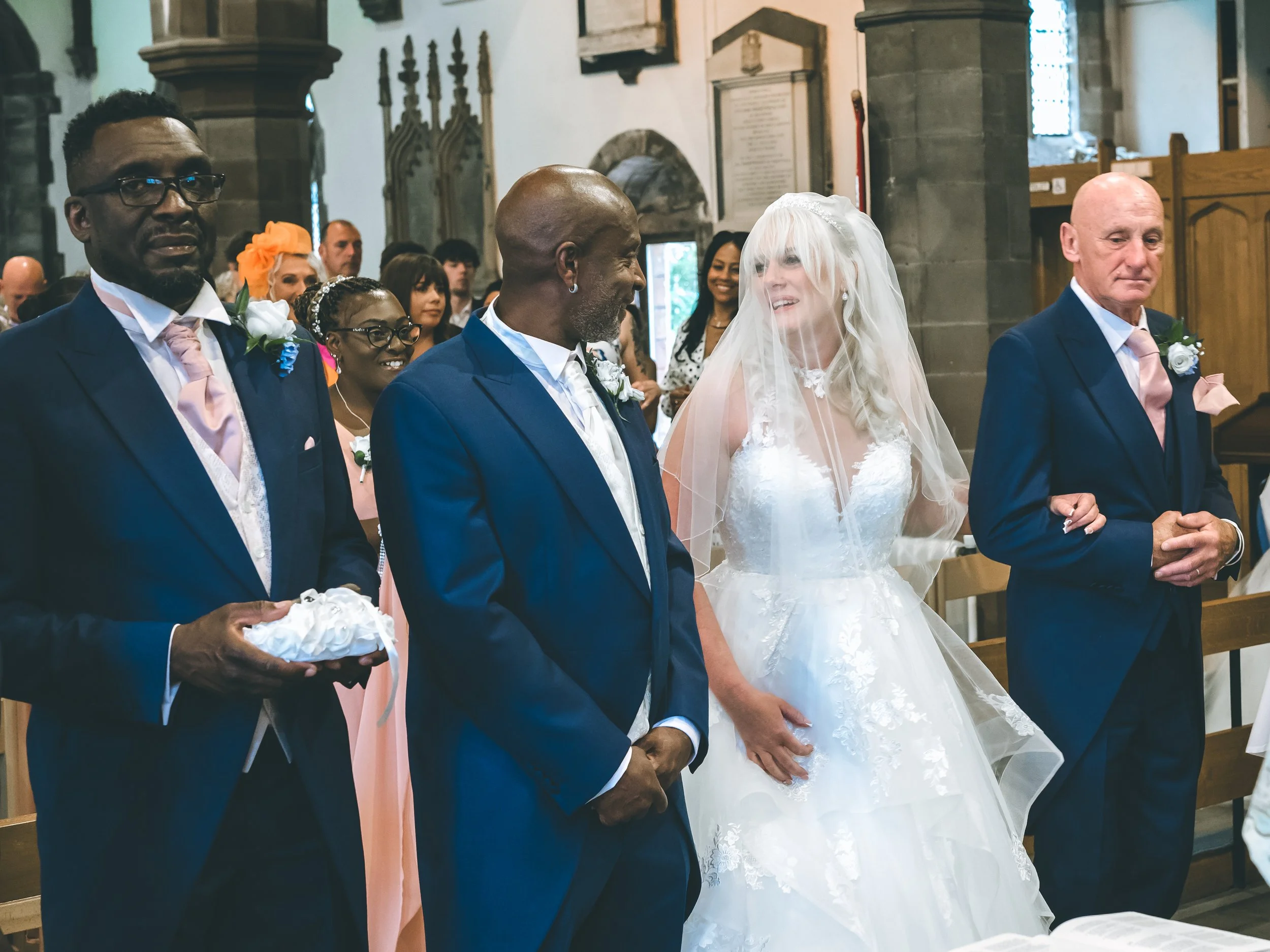 A bride and groom standing with their wedding party in a church during a wedding ceremony. The bride is wearing a white wedding dress and veil, smiling and looking at the groom. The groom is in a blue suit, holding hands with the bride, smiling. Surr