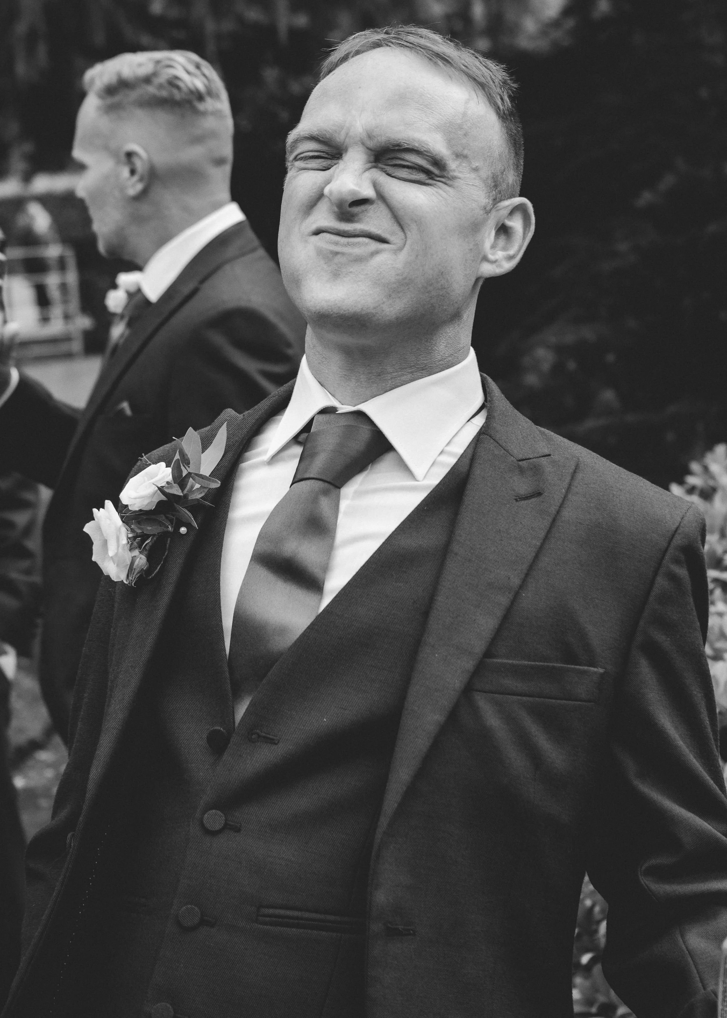 Black and white photo of two men dressed in formal suits, with the man in the foreground smiling with his eyes closed, and the man in the background looking to the side, both at an outdoor event.