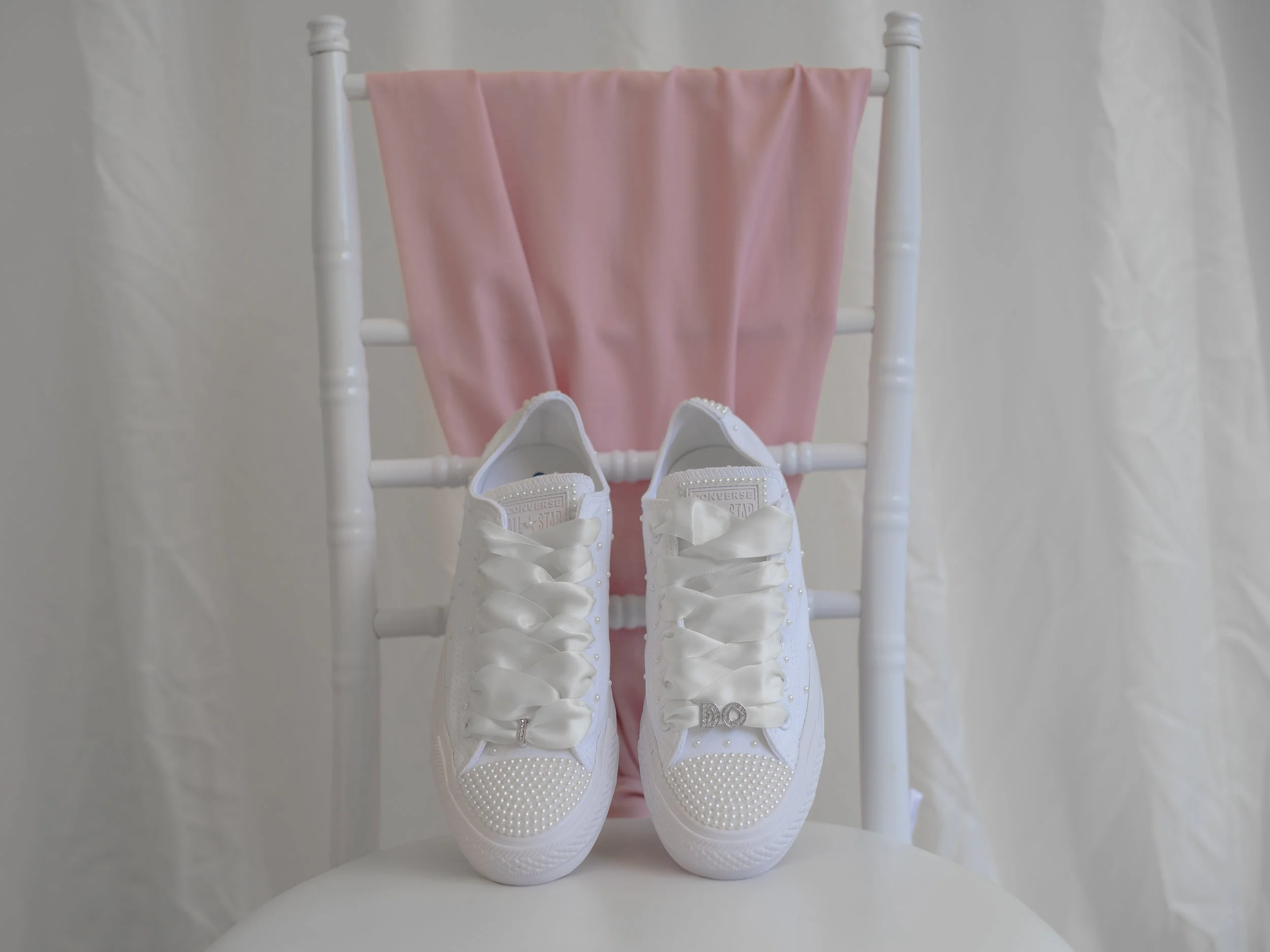 Pair of white sneakers with bows, pink clothing on a rack, and white shoes on a white surface.