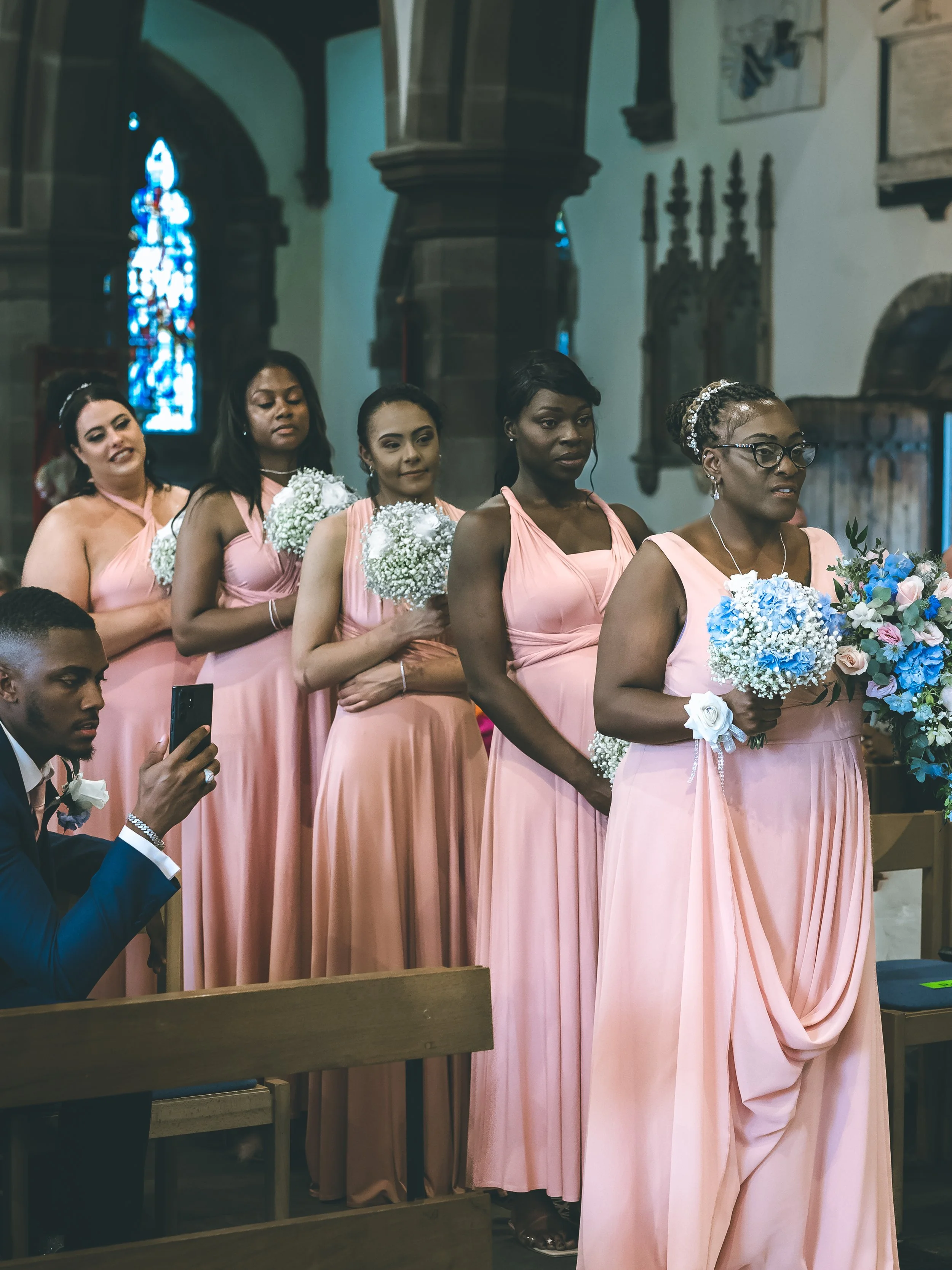 A group of women dressed in matching pink bridesmaid dresses holding bouquets of flowers inside a church during a wedding ceremony.