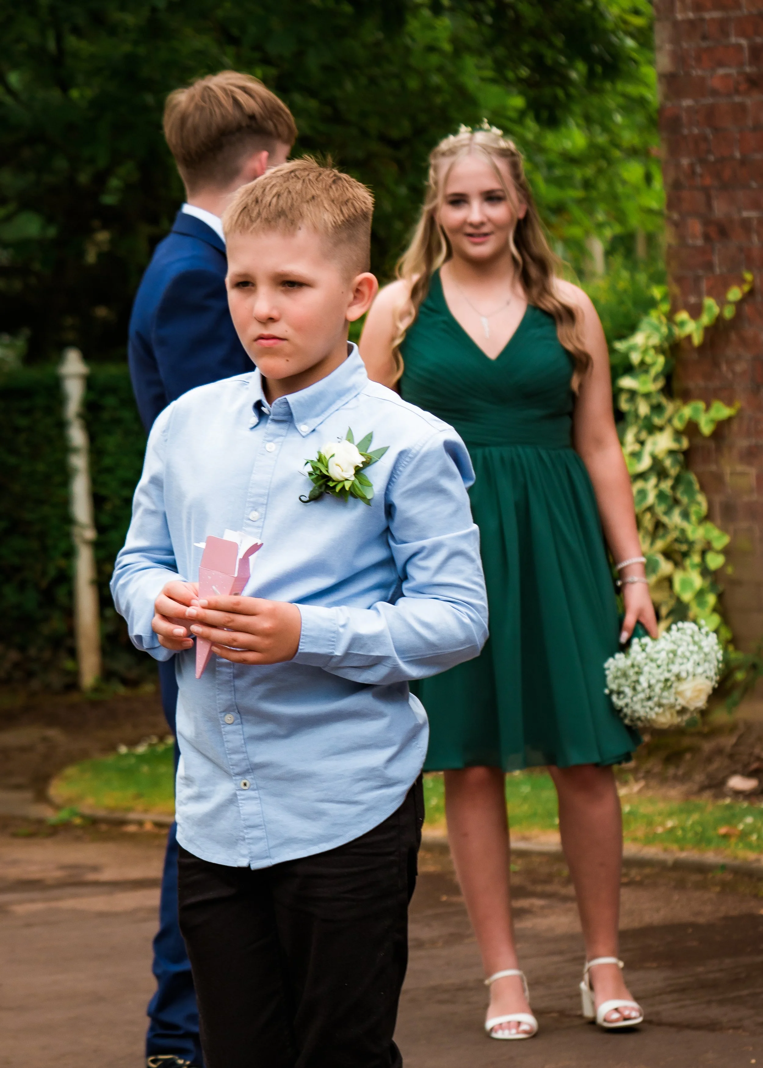 Young boy in a light blue dress shirt with a white boutonniere in a garden setting, holding a pink card, with two boys in suits and a woman in a dark green dress holding a bouquet of white flowers in the background.
