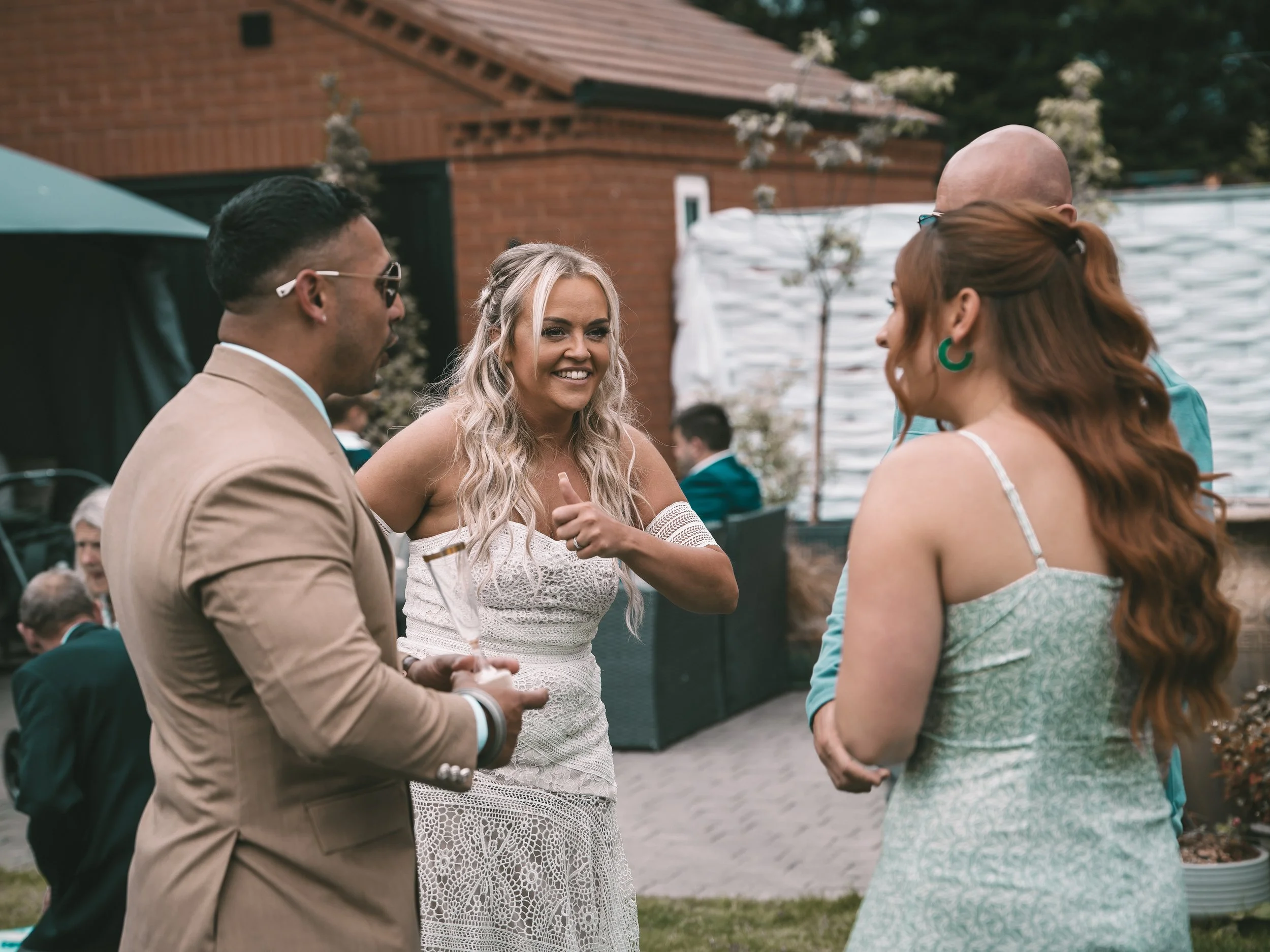 A group of four people, three men and one woman, standing outdoors in a garden or backyard setting, engaging in a conversation. They are dressed in semi-formal attire, with the woman in a white lace dress, and the men in suits or blazers. There is a 