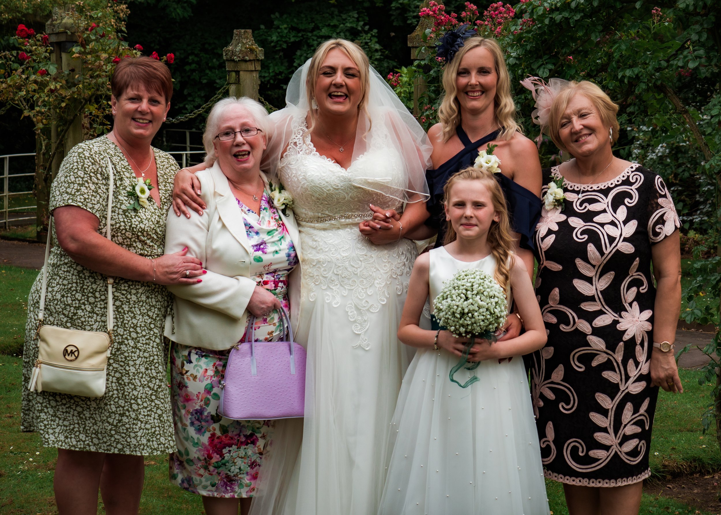 Group photo of women and a young girl at a wedding outdoors, with greenery and flowers in the background. The bride is in the center, wearing a white wedding dress and veil, holding a bouquet. Surrounding her are women dressed in colorful dresses, sm