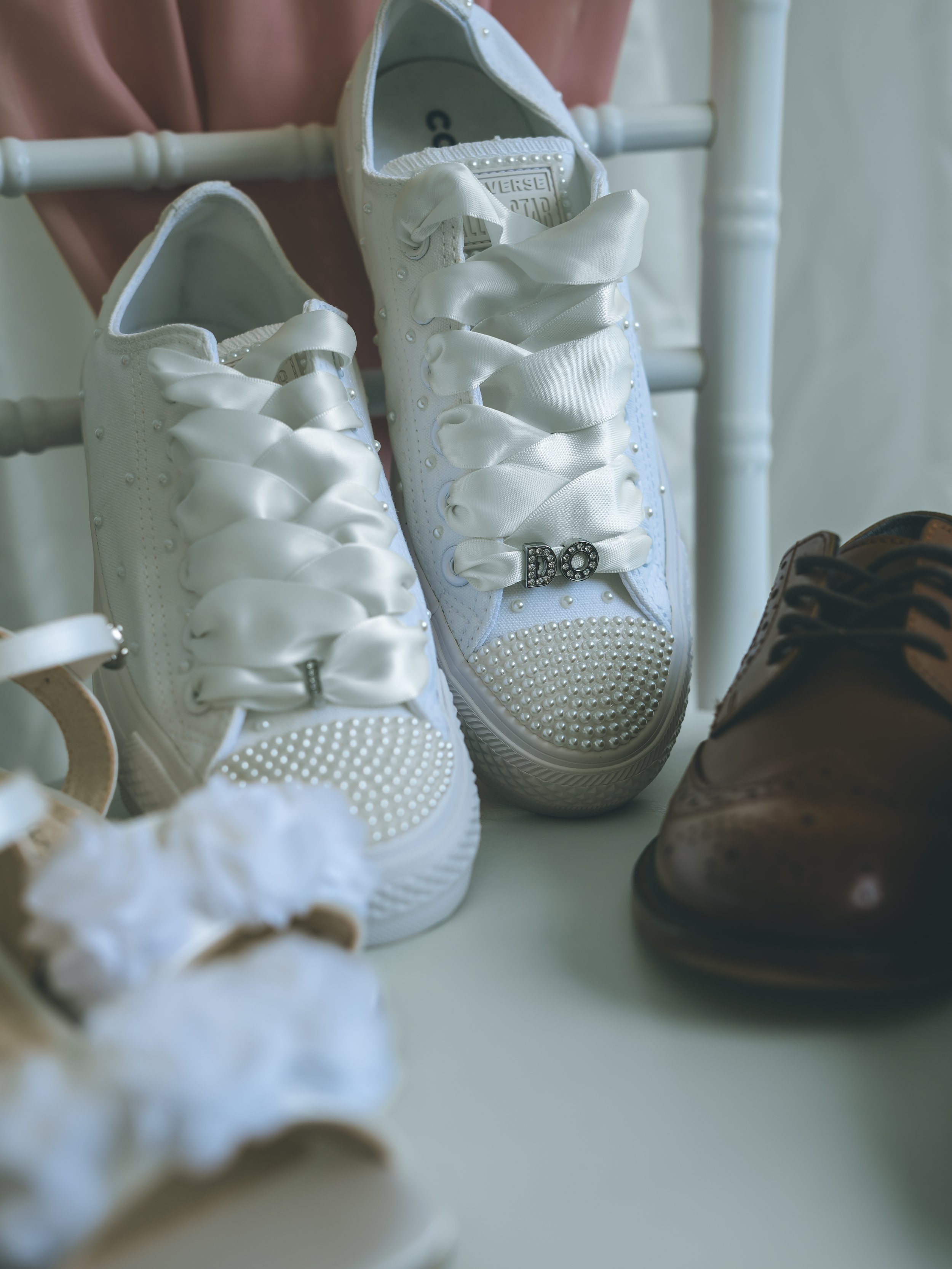 Pair of white sneakers with satin ribbons on a white surface, with brown shoes partially visible to the right and pink fabric in the background.