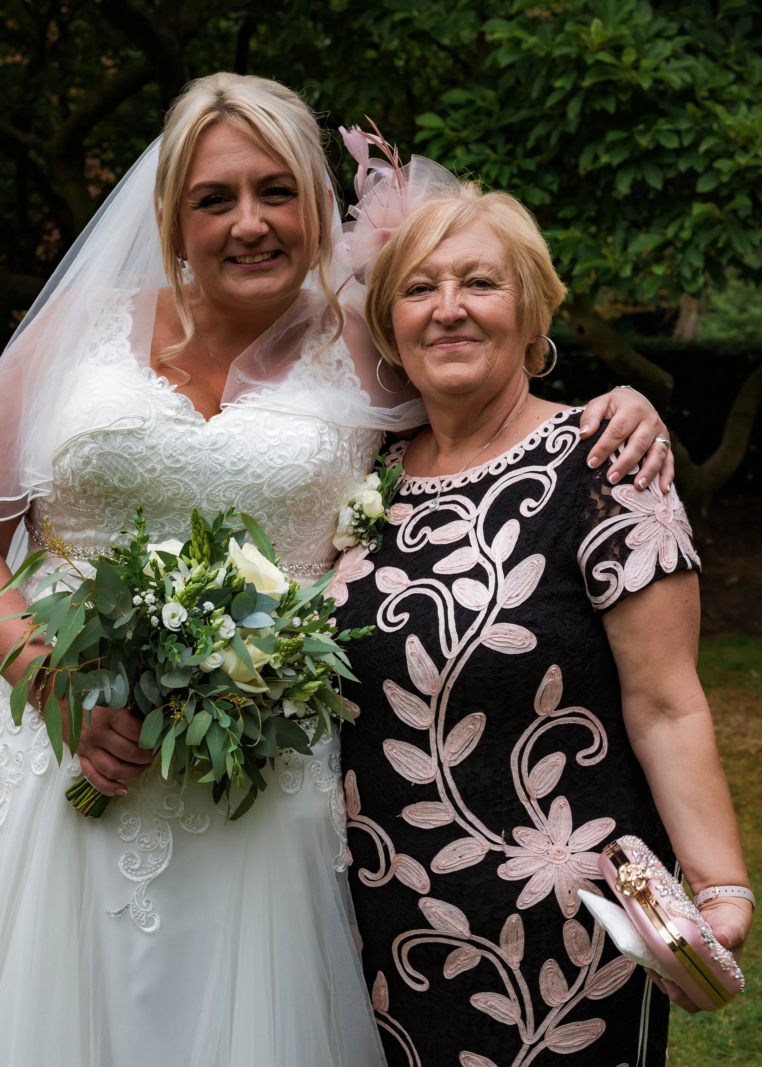 Two women at a wedding, one in a wedding dress holding a bouquet, the other in a black and pink floral dress, both smiling outdoors with greenery in the background.