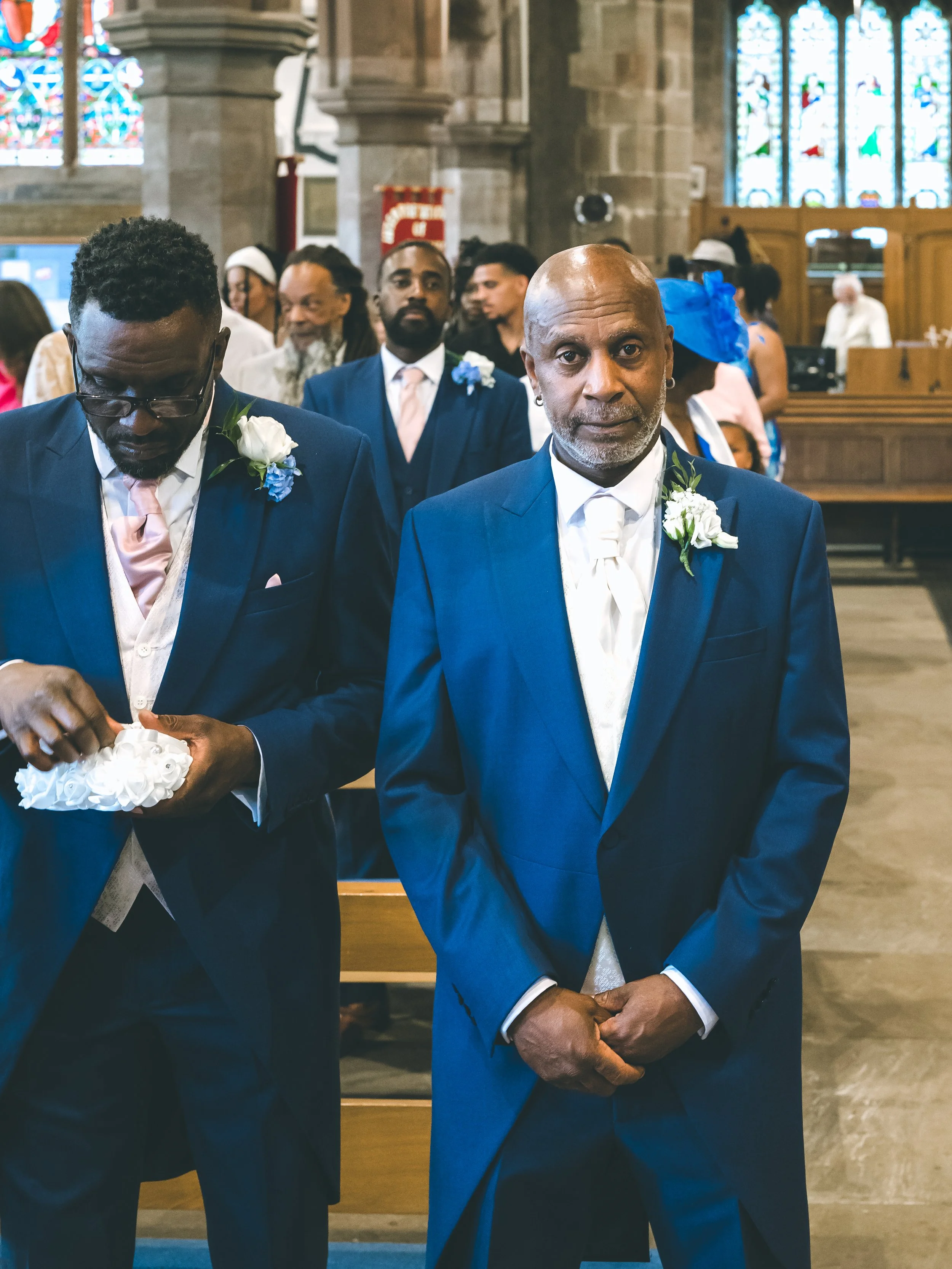 A group of men in suits with boutonnières attending a formal event in a church. The man in front has a bowtie, a boutonnière, and is standing with his hands clasped, looking at the camera. The church has stained glass windows and wooden pews.