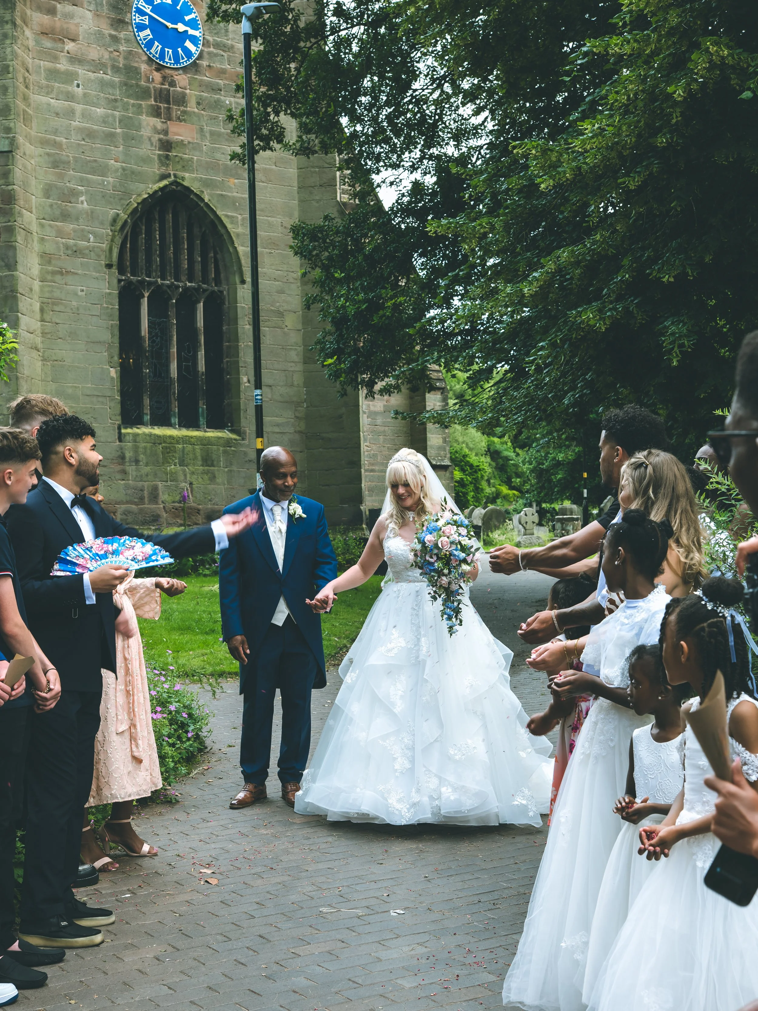A wedding ceremony taking place outdoors in front of a church with a stone facade and a large clock. The bride and groom are holding hands, surrounded by guests and children in white dresses. The bride is wearing a white wedding gown and holding a bo