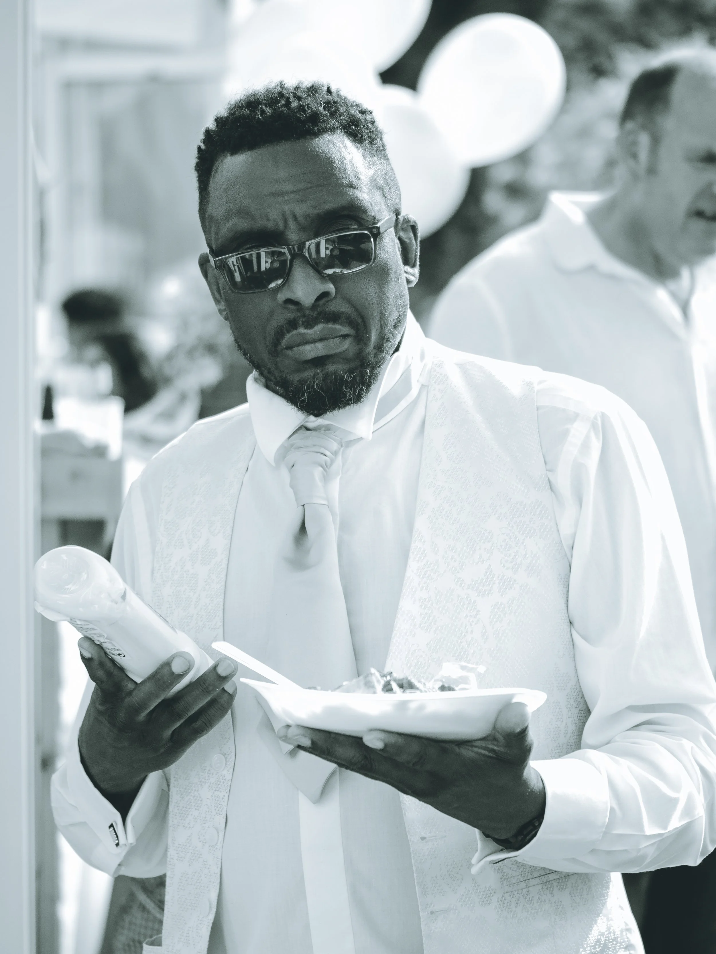 Man in formal attire wearing sunglasses, looking at his phone, holding a tray with food, outdoors, with balloons in the background, black and white photo.