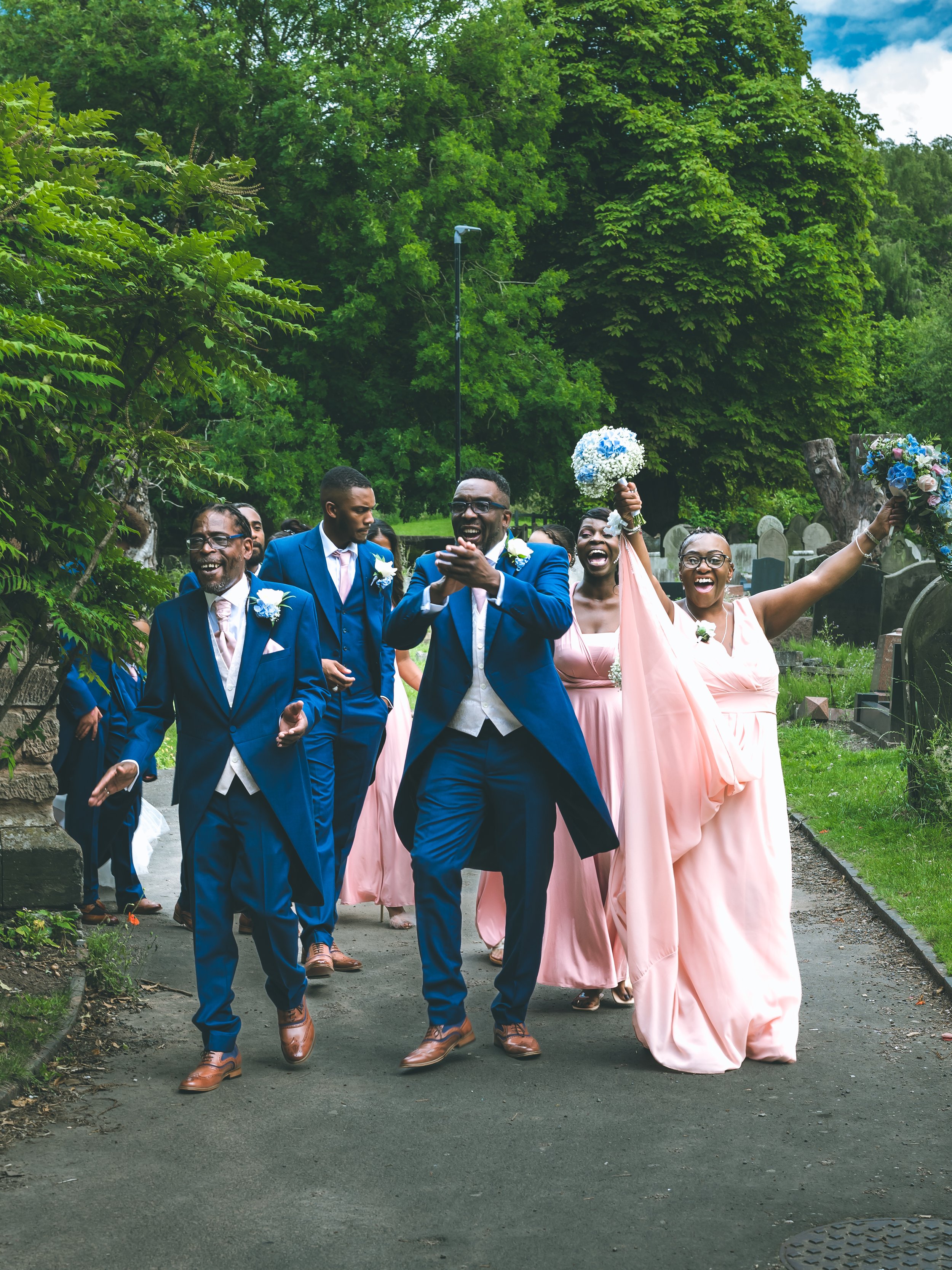 A group of people at a wedding walk outdoors on a path lined with trees, wearing formal attire, with some holding bouquets and smiles.