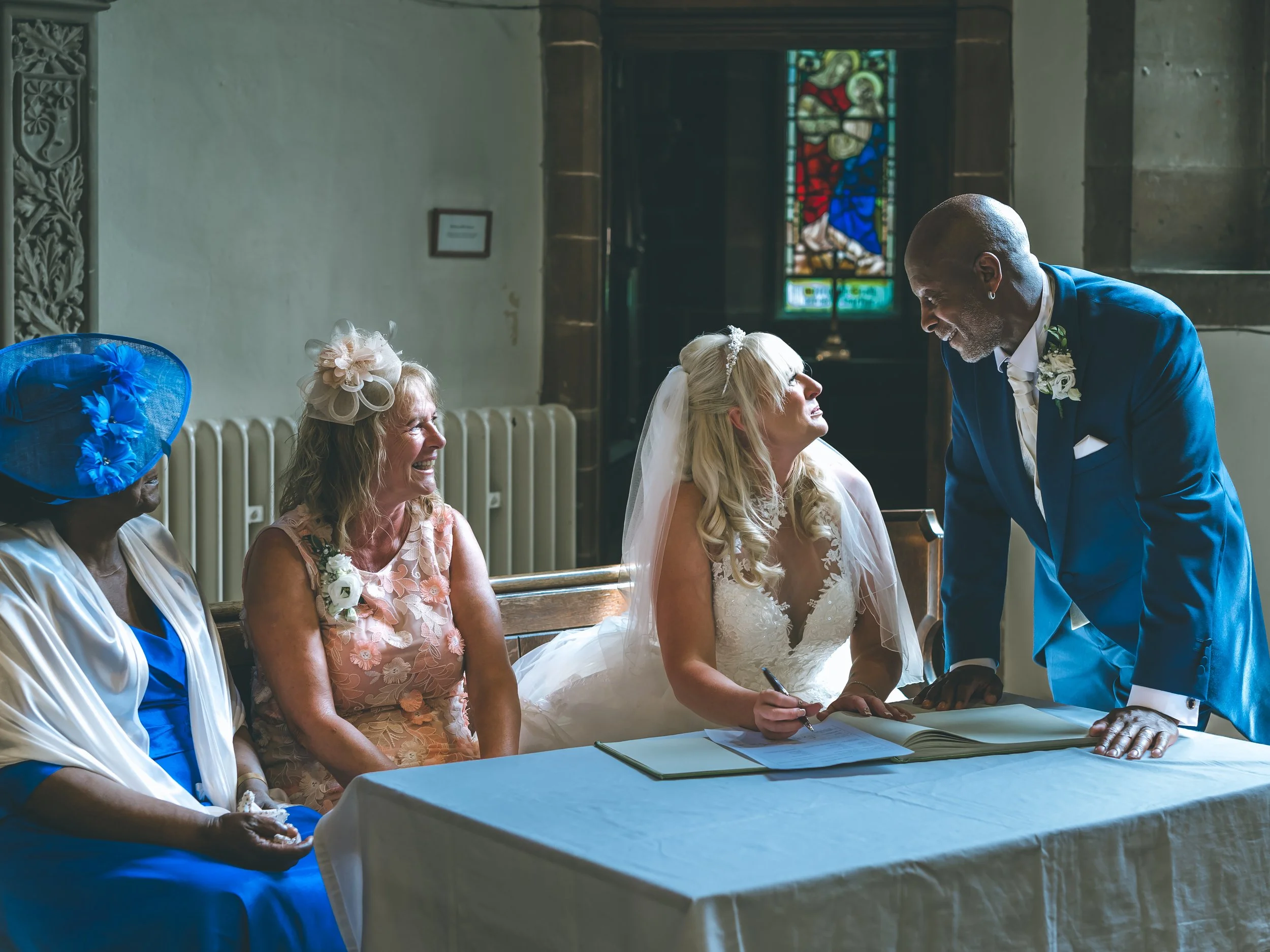 A bride signing a marriage register at the altar during her wedding ceremony with a man standing beside her. Other women, including one in a pink dress and blue hat, are seated nearby in a church with stained glass windows in the background.