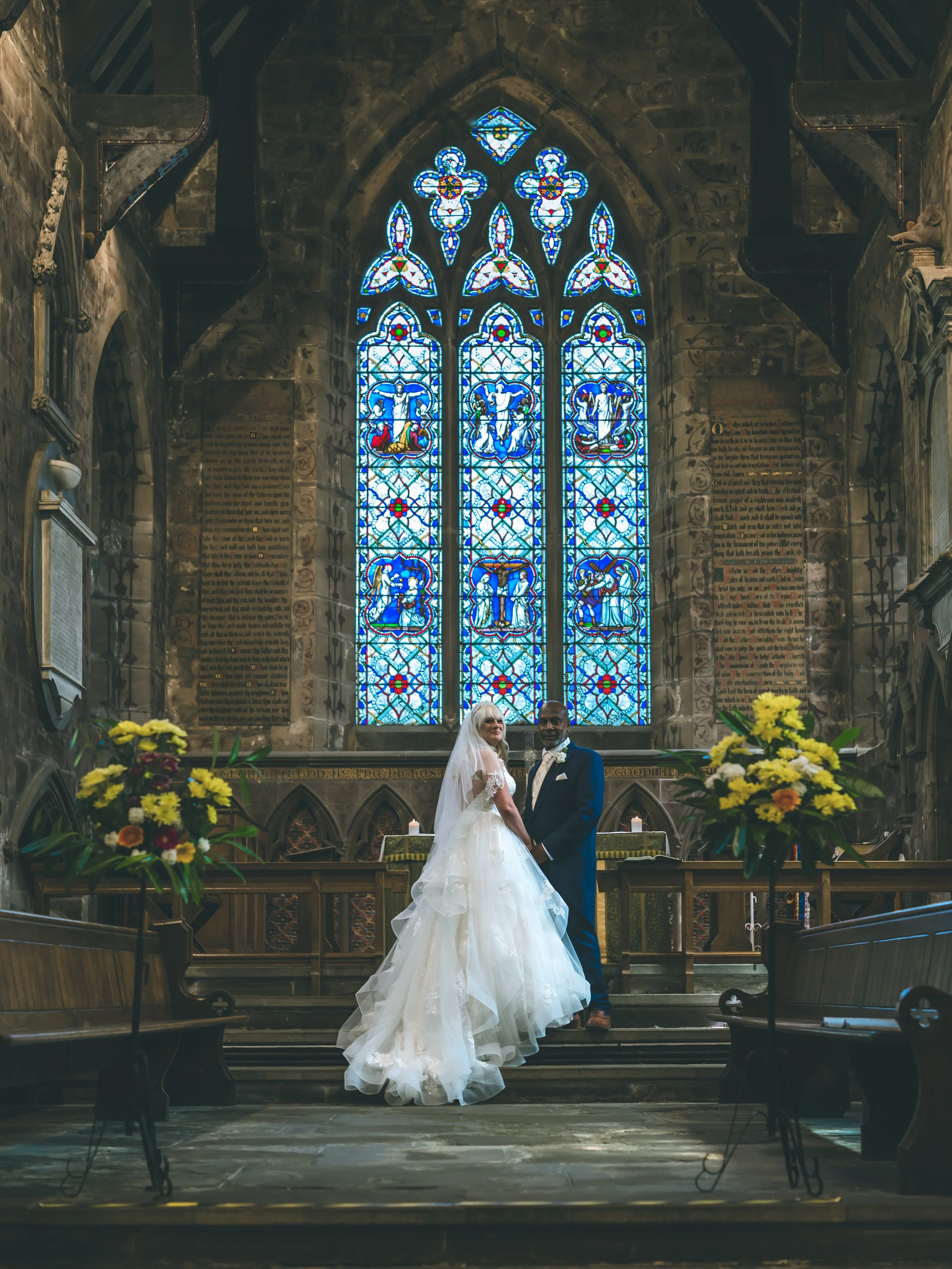 A bride and groom standing in a church during their wedding ceremony, with stained glass windows and floral arrangements in the background.