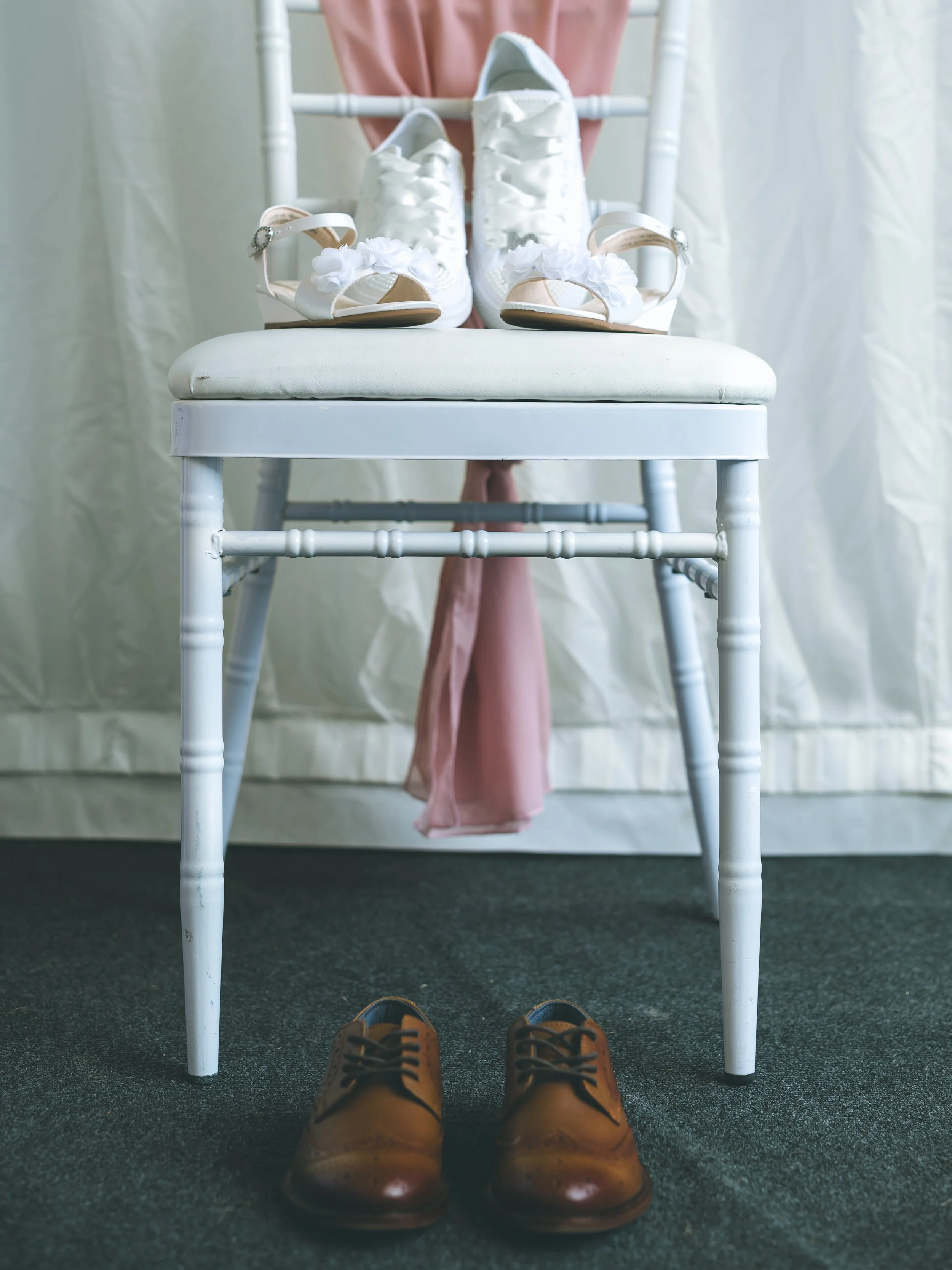 A pair of brown dress shoes on a dark carpet. The perspective is from the shoes, looking up at a white chair with white sneakers, pink heels, and other shoes on it, with a person's pink dress and white curtain in the background.