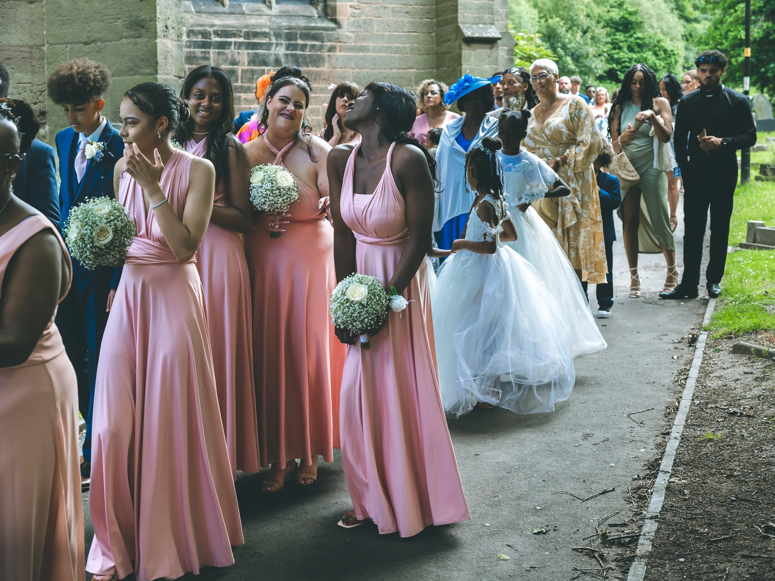 A group of people dressed for a wedding, including bridesmaids in pink dresses holding bouquets of white roses and baby's breath, gathered outside near an old stone building with lush greenery in the background.