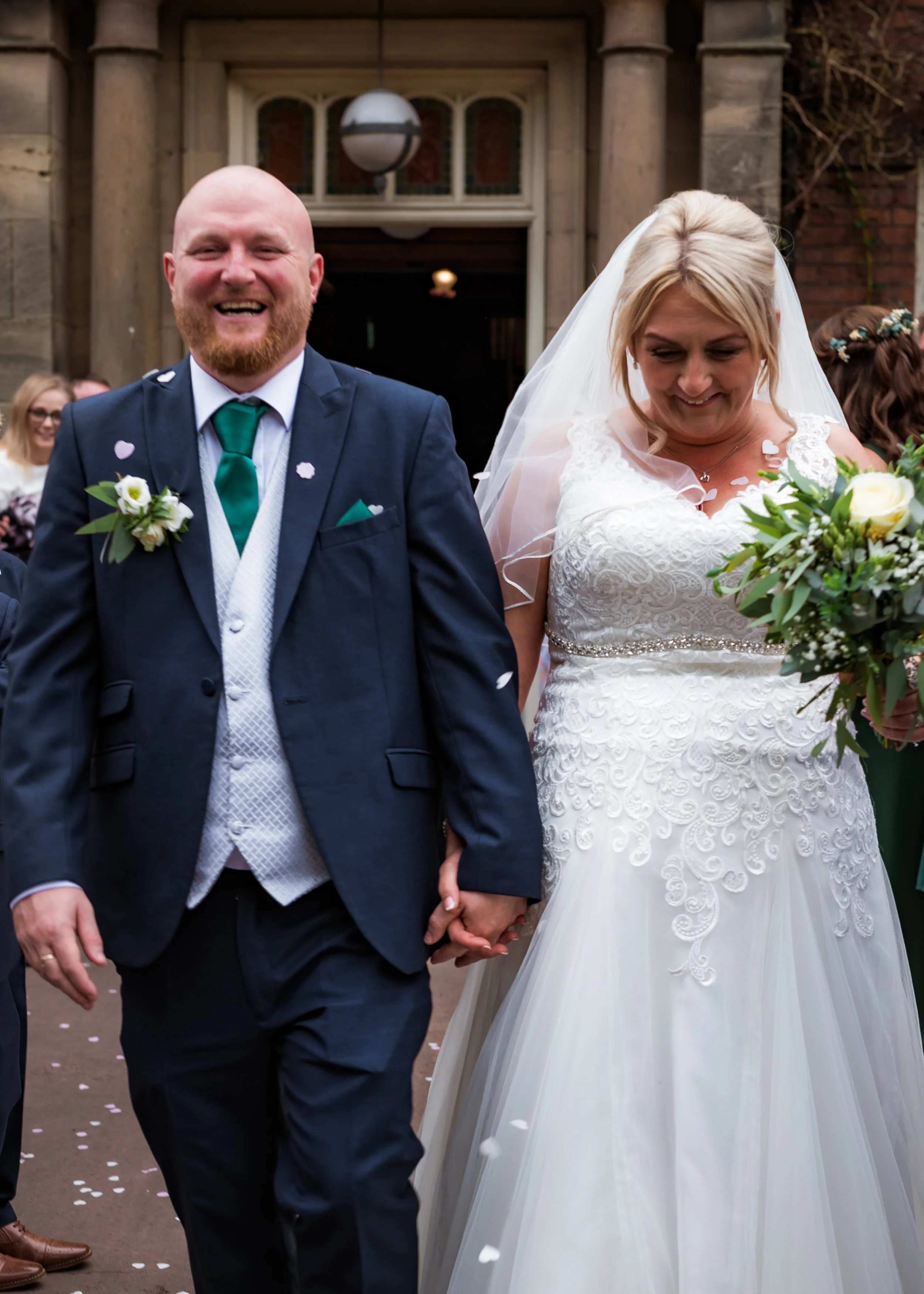 A newlywed couple holding hands and walking outside a church, the groom is smiling and wearing a navy suit with a green tie, and the bride is looking down with a gentle expression, wearing a white lace wedding gown and veil, surrounded by wedding gue