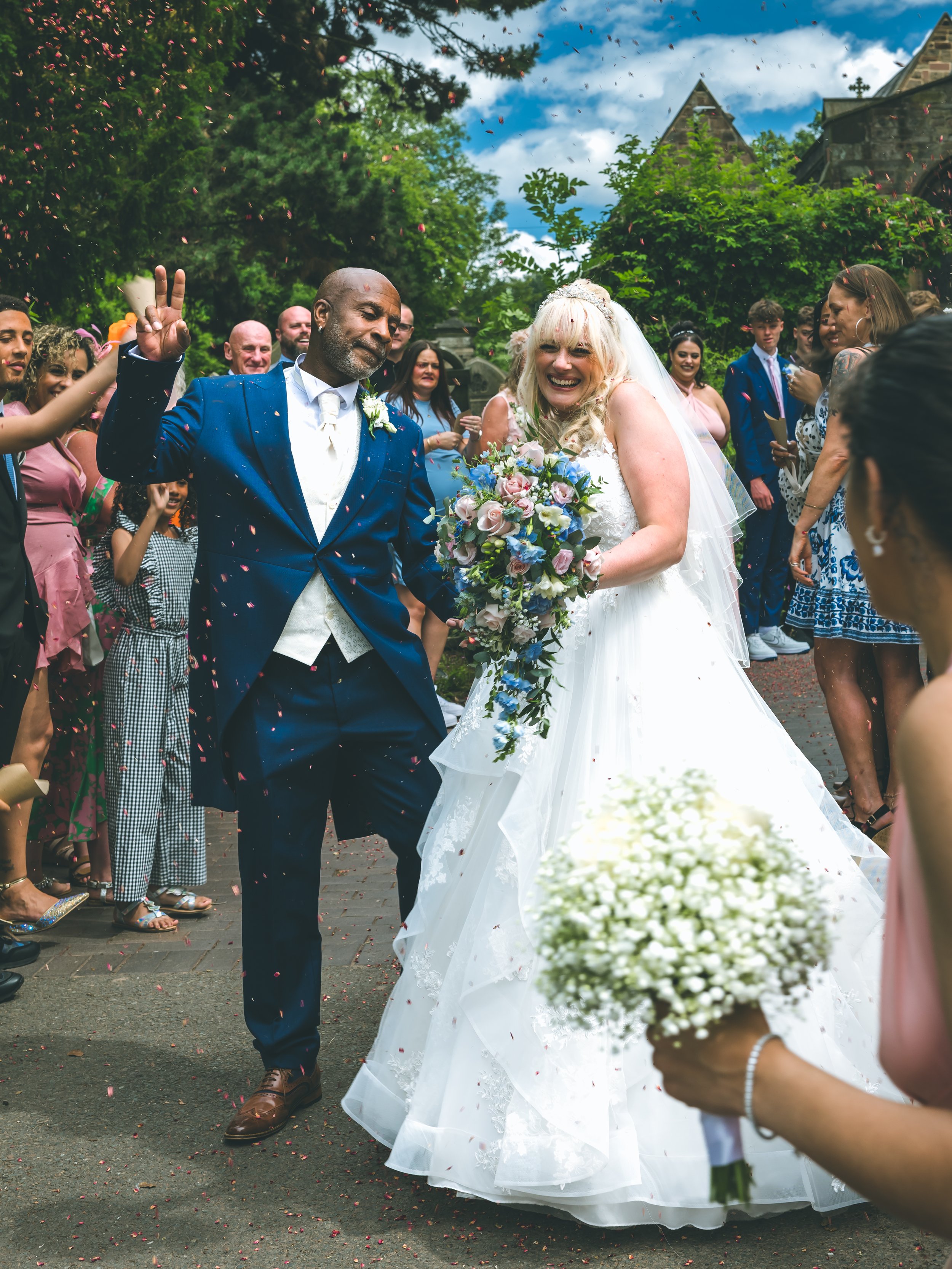 A wedding celebration outdoors with a bride and groom walking together, surrounded by guests showering them with confetti, and greenery in the background.
