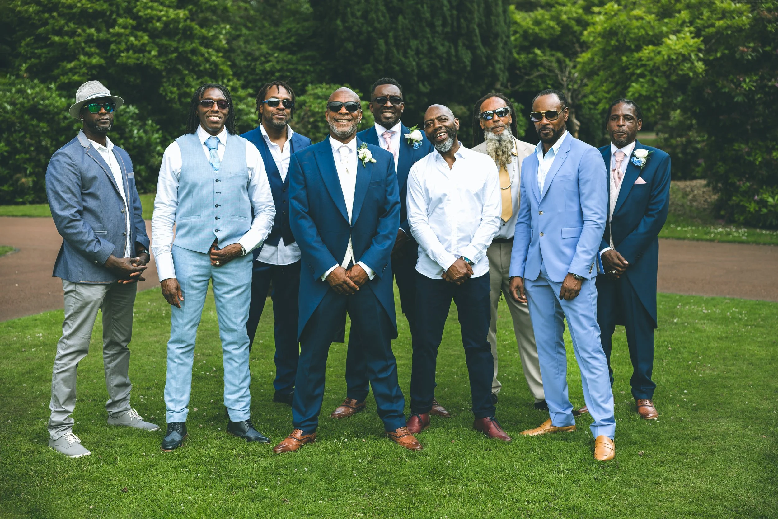 A group of eleven men dressed in formal suits, some wearing sunglasses, standing outdoors on grass with a backdrop of trees, smiling for a photo.