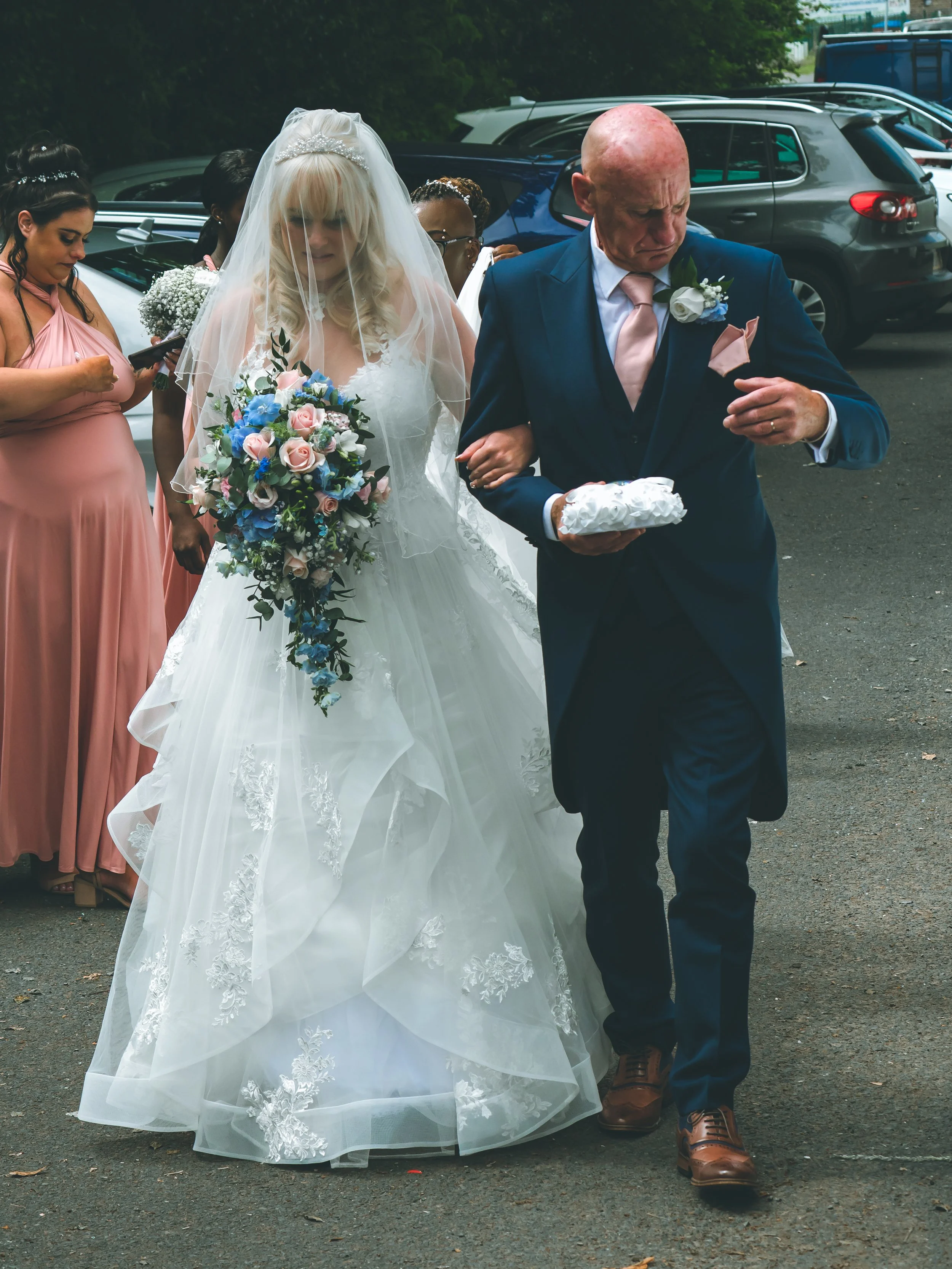 A bride in a wedding dress and veil walking arm-in-arm with an older man in a blue suit and pink tie, likely her father, at a wedding. Women in pink dresses are seen behind them, one using her phone. They are outdoors, near parked cars.
