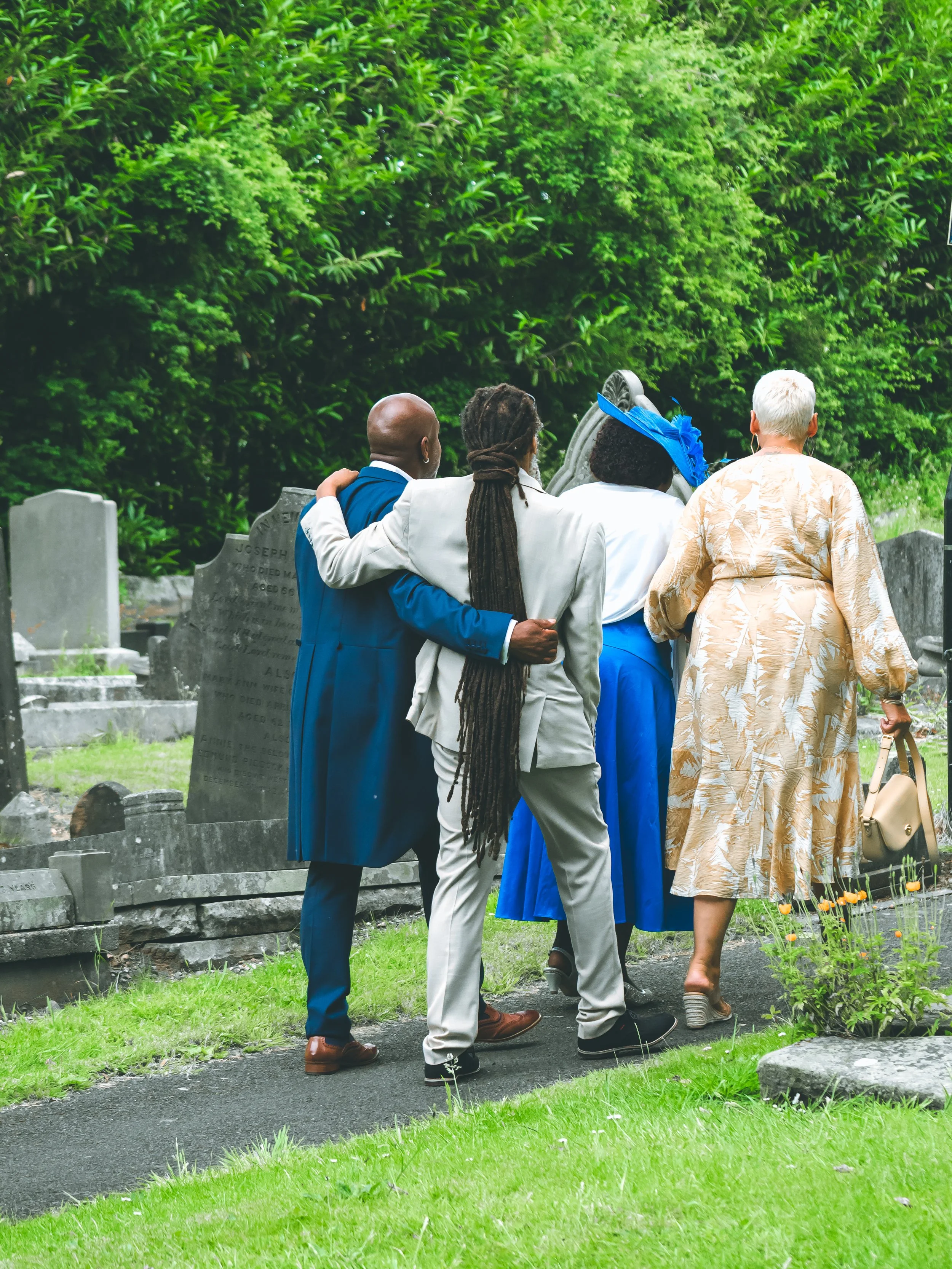 Four people dressed in formal attire walking together in a cemetery, with headstones and green trees in the background, and one person carrying a beige purse.