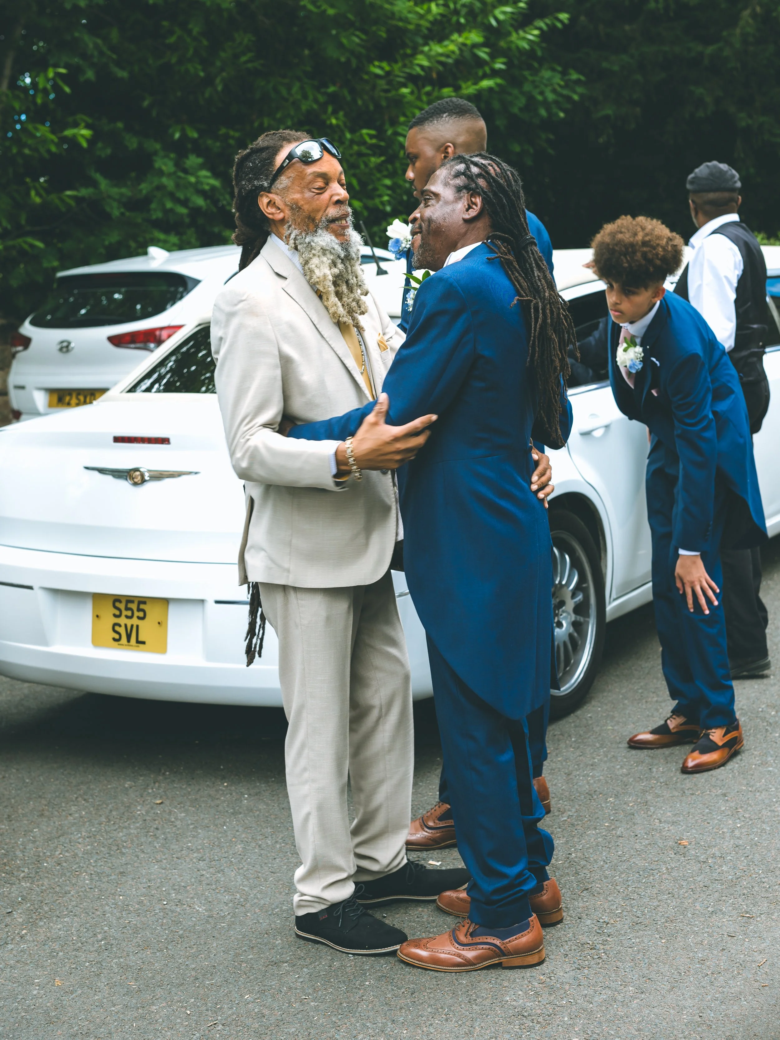 Two men are smiling and holding each other's hands in an outdoor setting with cars and other people in the background.