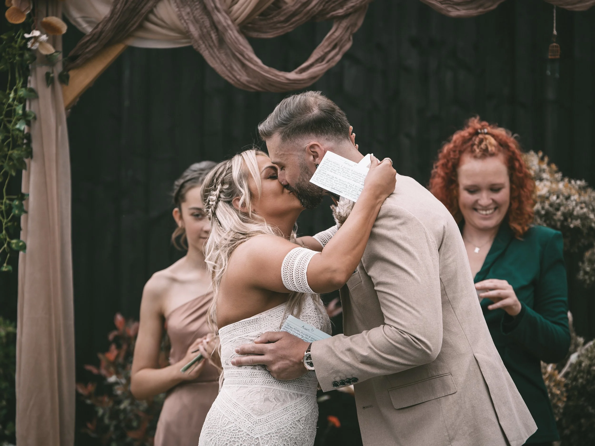 A couple is kissing at their wedding ceremony, with the bride holding a handwritten note and both dressed in wedding attire. Friends and family are smiling in the background.