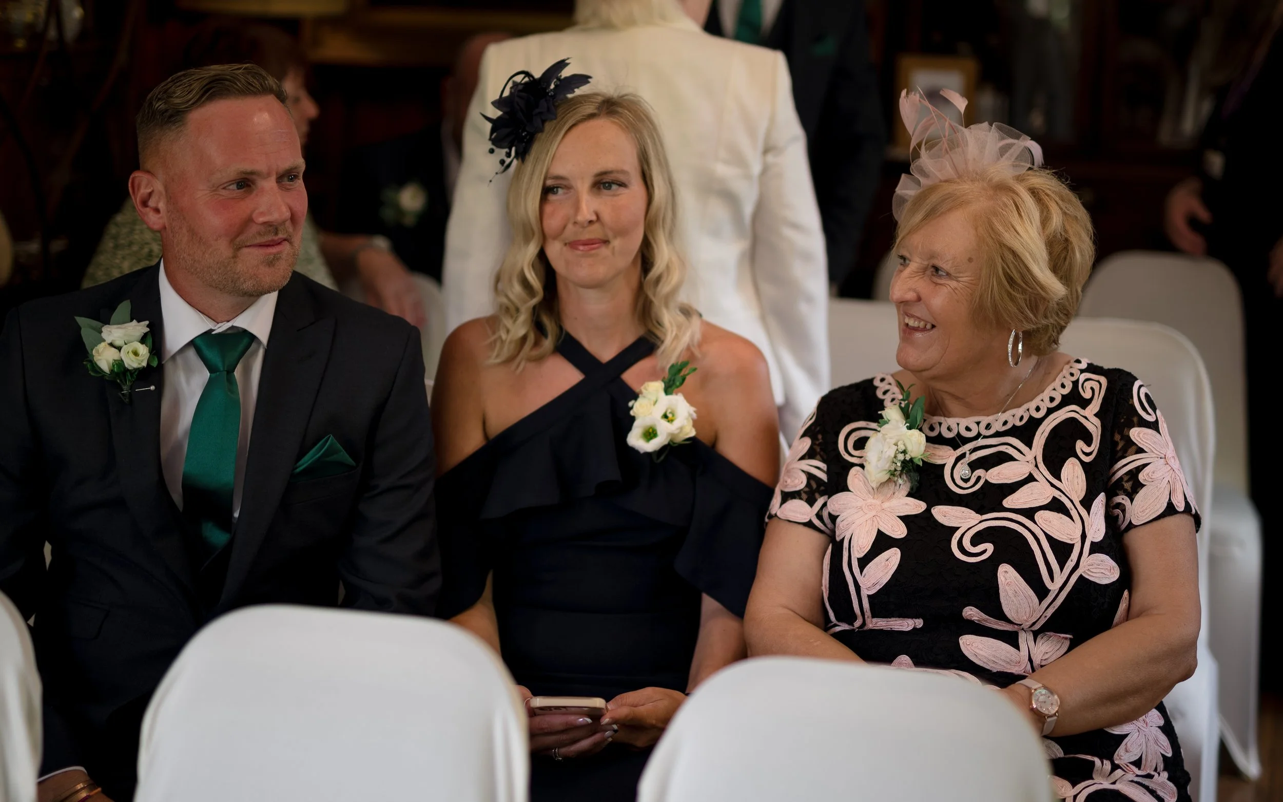 Three women sitting at an event, dressed in formal attire, wearing corsages, and smiling, with a man in a suit partially visible on the left.