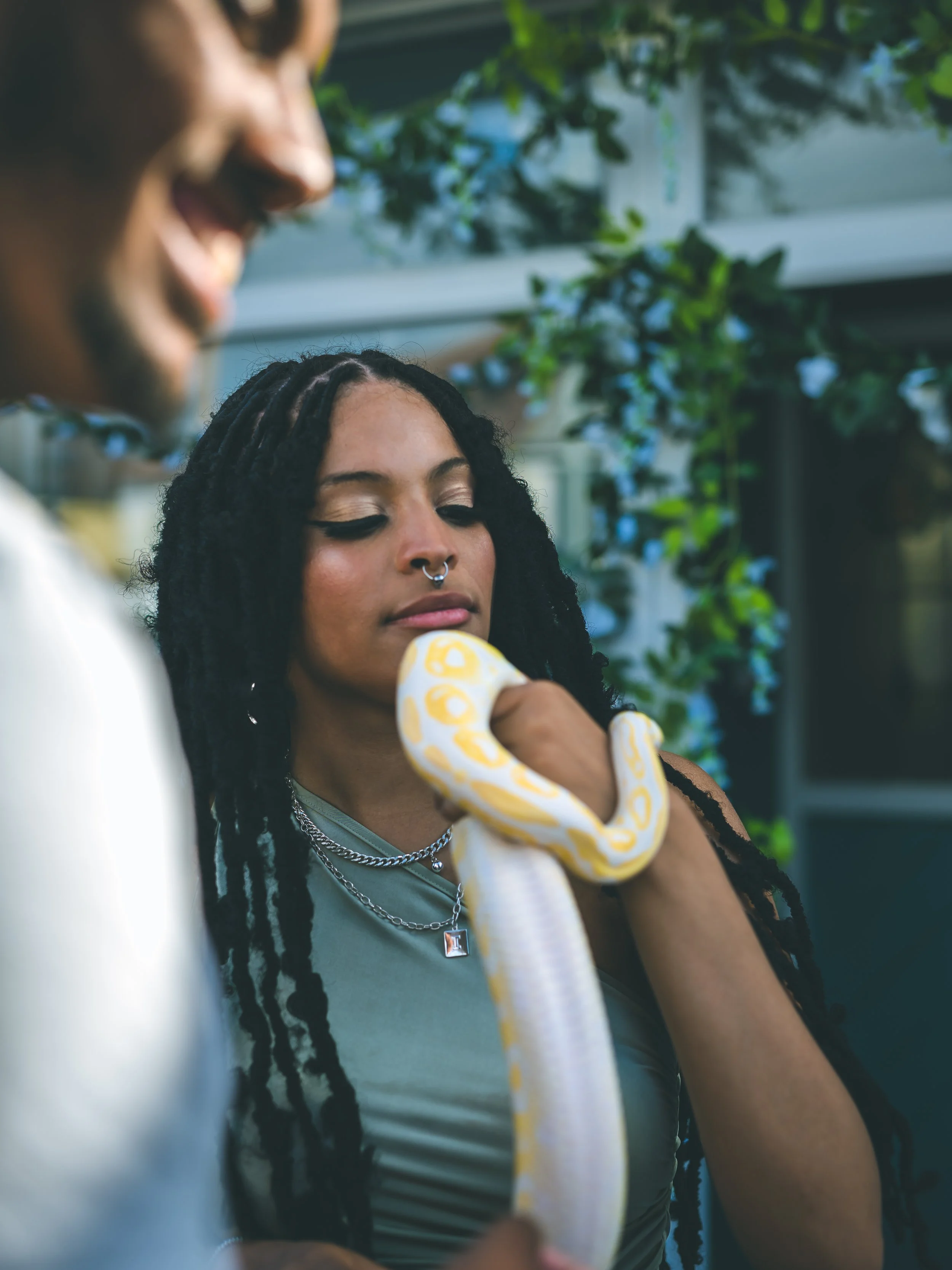 A woman with dreadlocks and a septum piercing looking at a yellow snake that is wrapped around her arm, with another person partially visible in the foreground, outdoors with greenery and a house in the background.