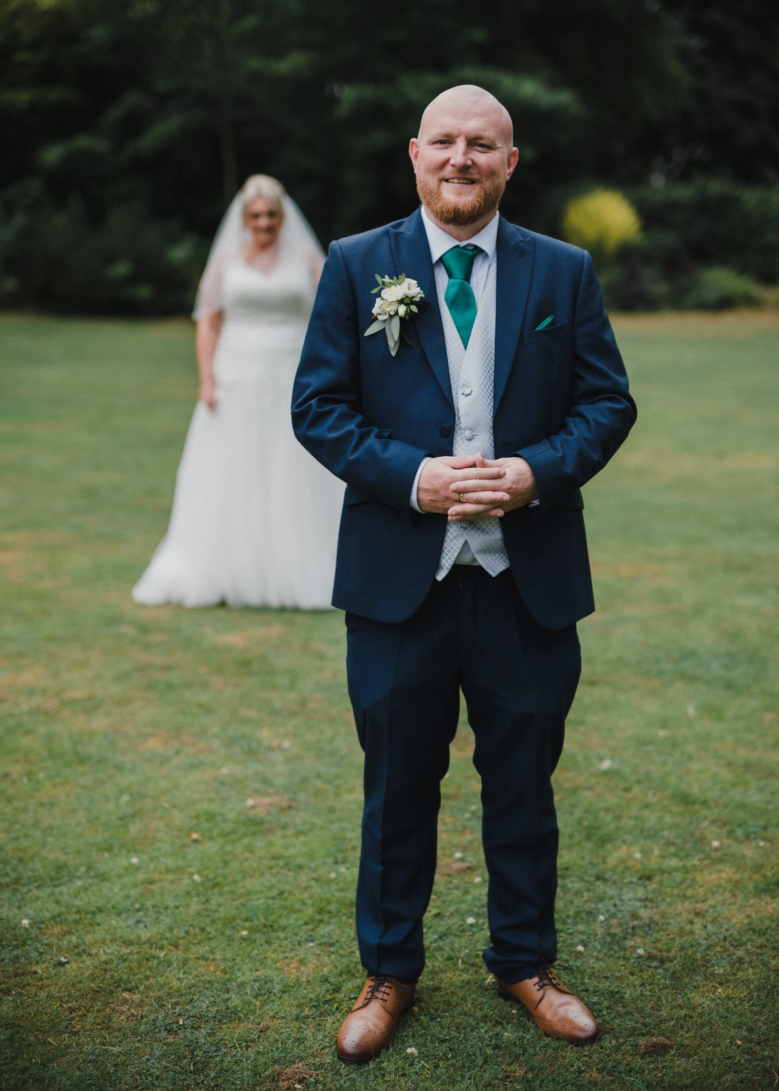 A man in a dark blue suit, white shirt, teal tie, and tan shoes standing outdoors on grass, smiling at the camera, with a woman in a white dress and veil in the background at a wedding.