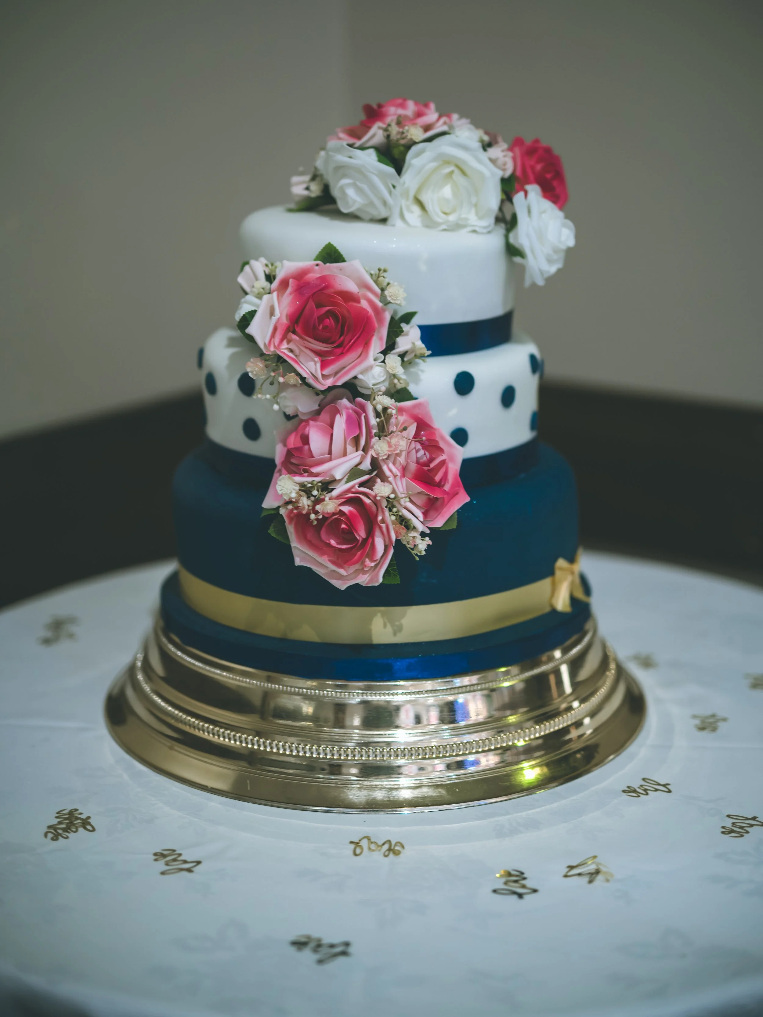 A two-tiered wedding cake decorated with white, pink, and red roses, positioned on a gold cake stand with a white wrapping paper printed with gold lettering underneath.