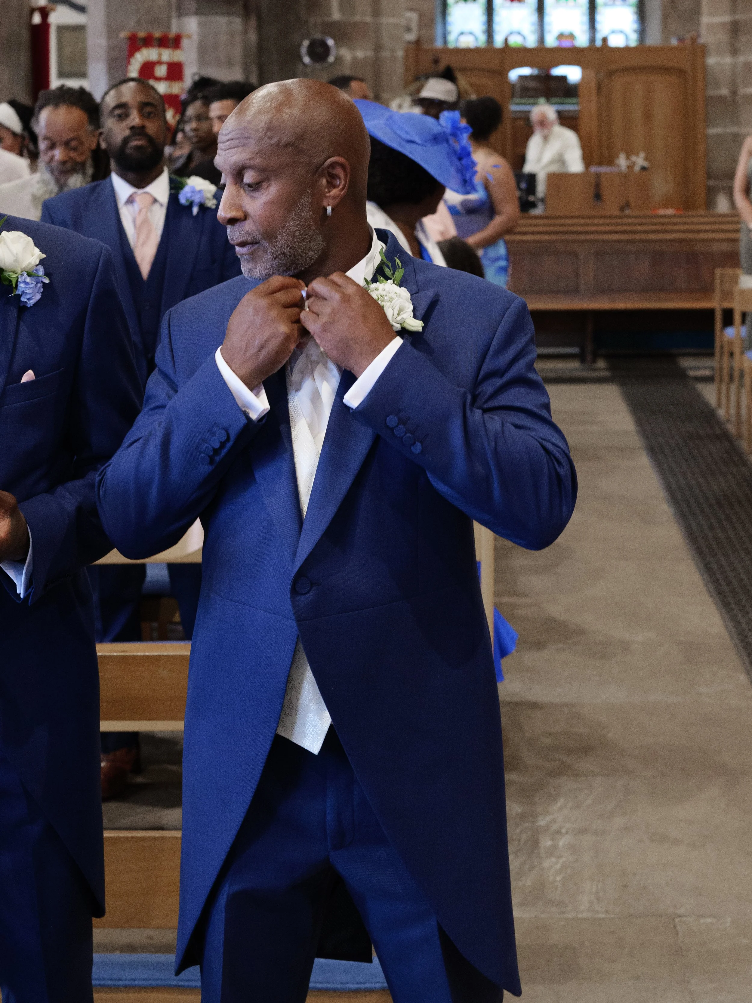 A man in a blue suit is adjusting his tie inside a church during a ceremony. Other people are standing behind him, dressed in formal attire, some with flower boutonnieres. The background shows stained glass windows and church furnishings.