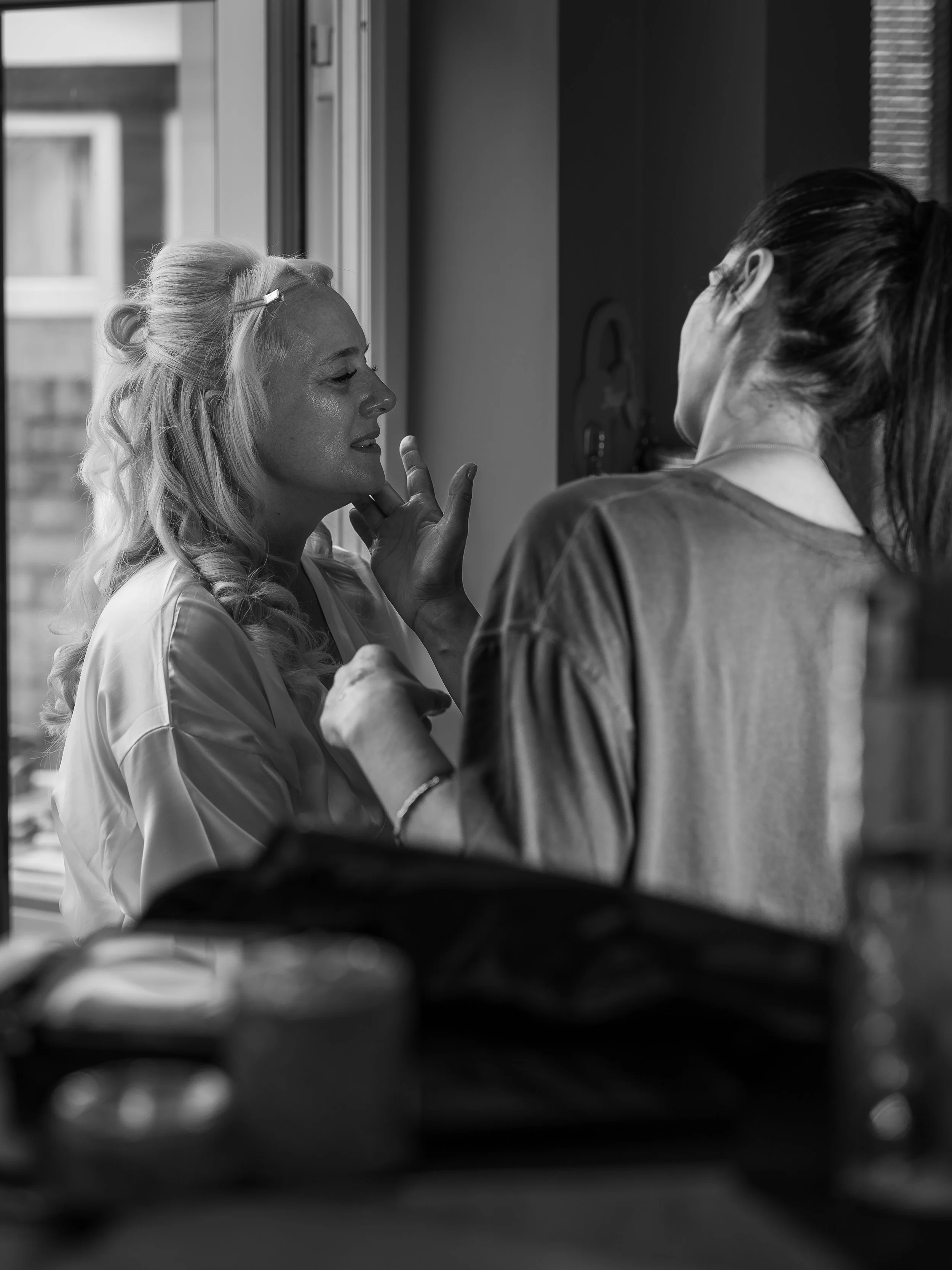 A woman with blonde hair styled in loose curls and a hair clip, smiling and touching her face, is seen through a window. Another woman with dark hair tied back, facing her, is also visible. The scene occurs indoors near the window, with city building