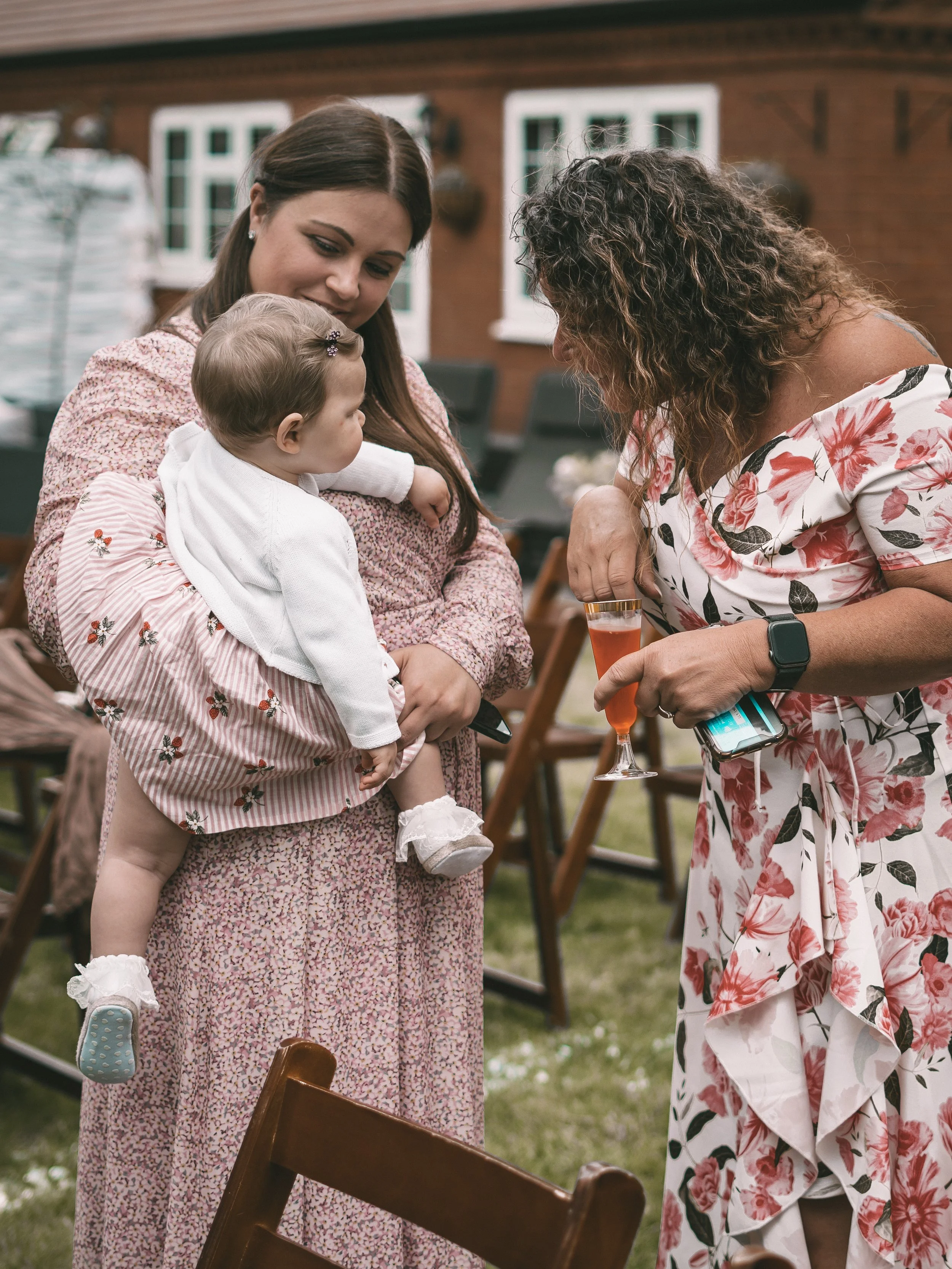 A woman holding a young girl in her arms while another woman with curly hair leans forward holding a drink at an outdoor gathering.