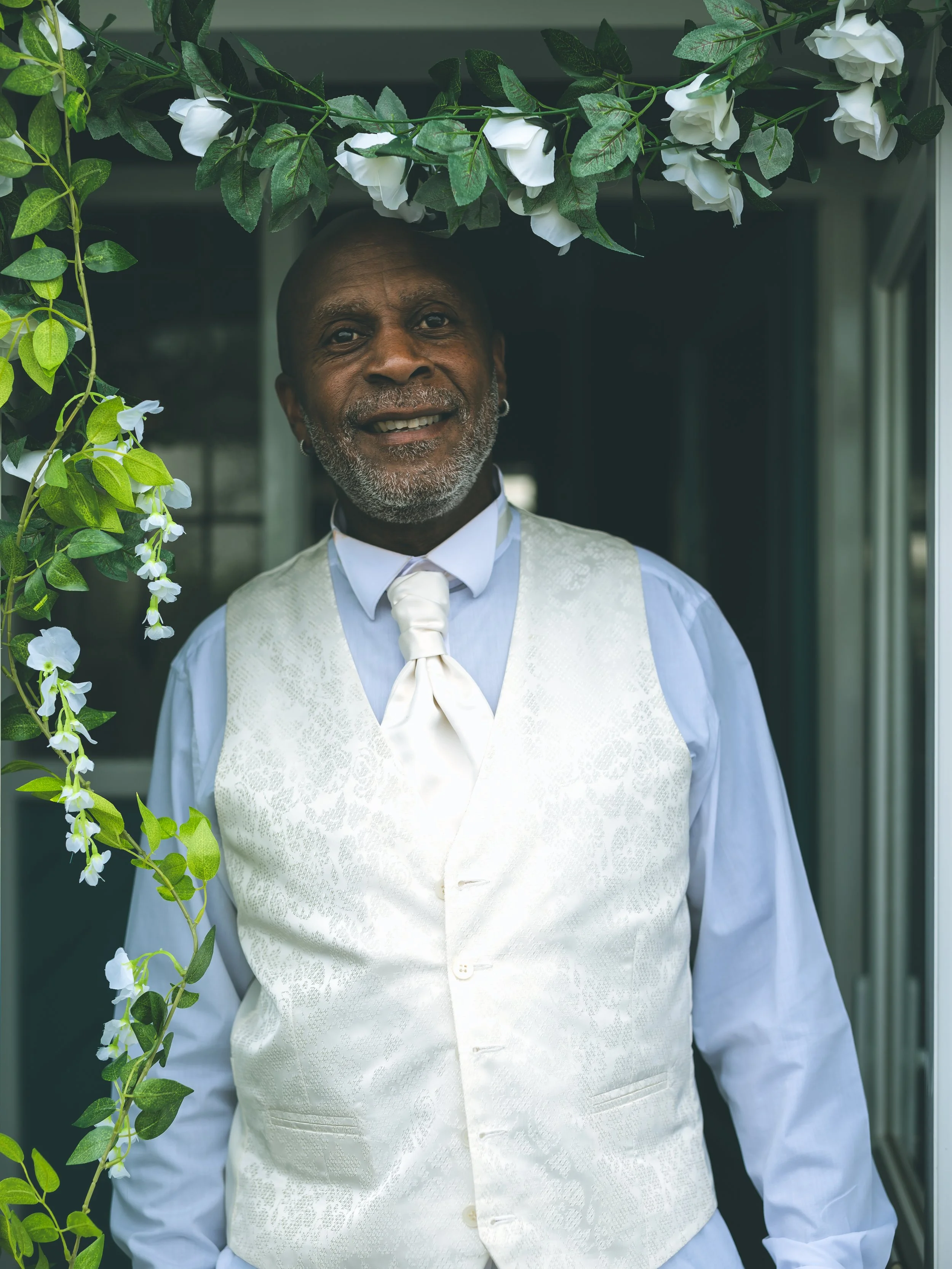 A smiling man dressed in a white vest, shirt, and tie, standing outdoors near greenery and white flowers.