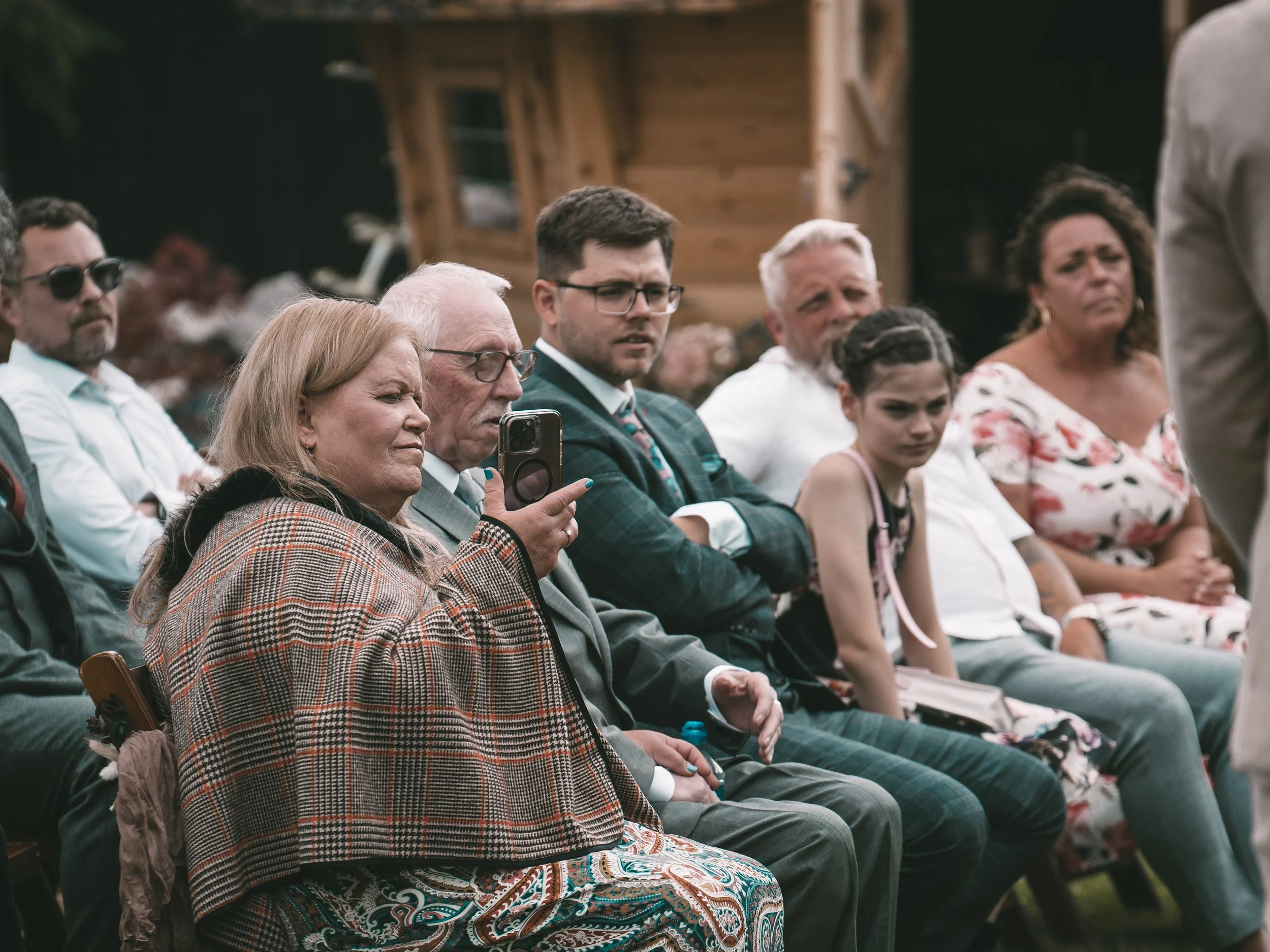 A group of people sitting outdoors, attending an event or gathering. The group includes men and women of different ages, some wearing glasses, with a wooden building in the background.