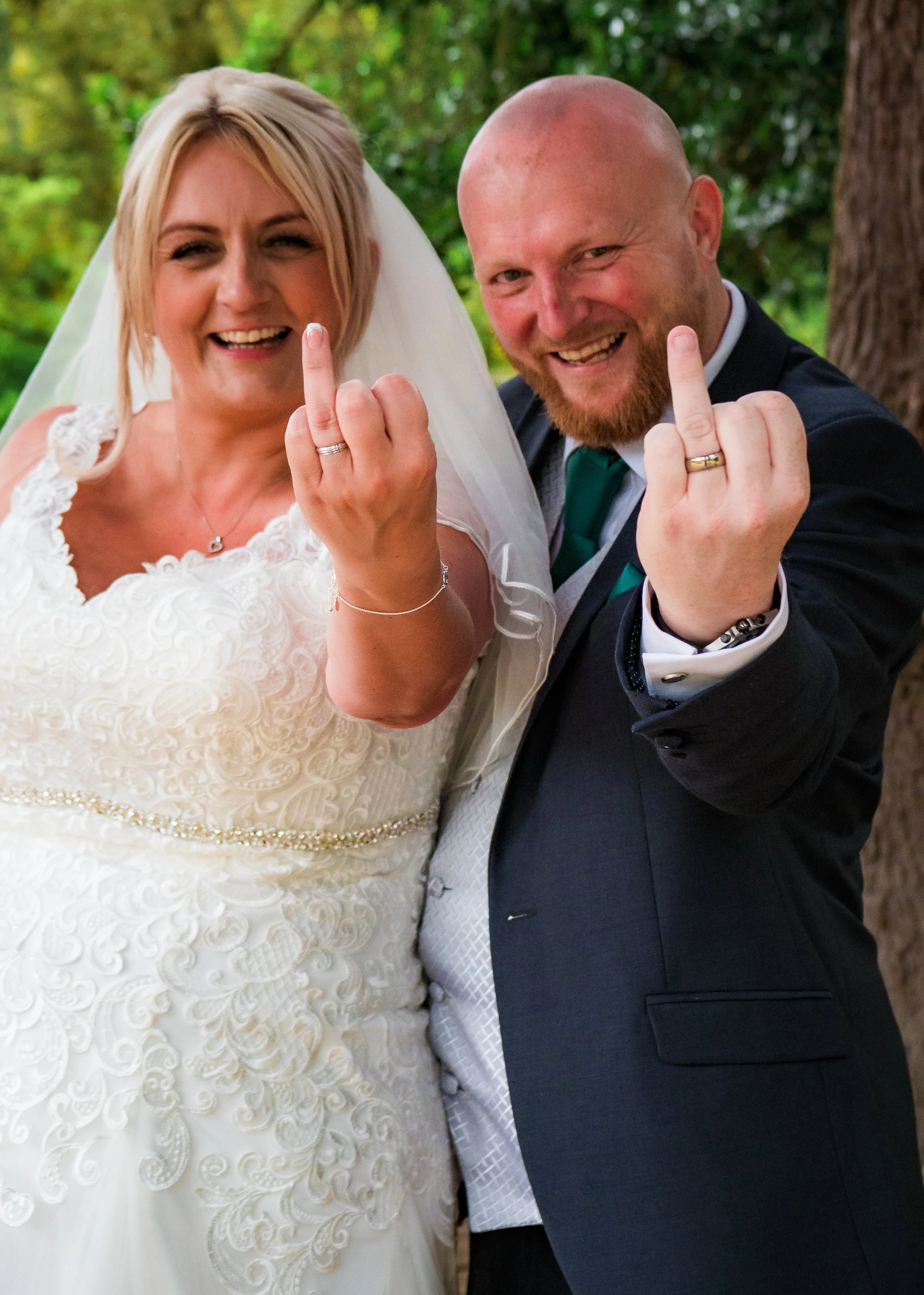 A newlywed couple, the bride in a white lace wedding gown with a veil and the groom in a dark suit, both smiling and showing their middle fingers as a gesture of defiance, outdoors with green foliage in the background.