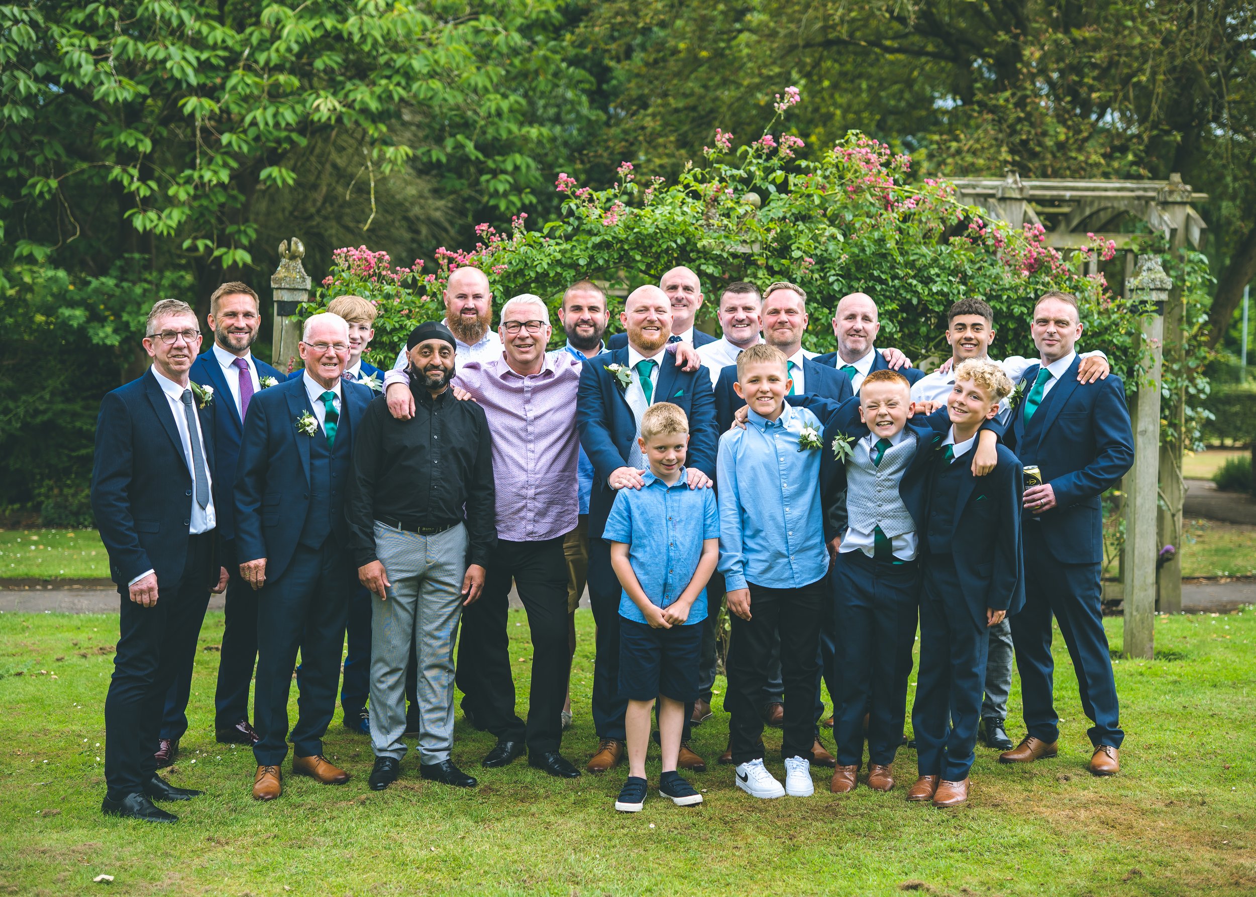 A group of men and boys dressed in formal attire, including suits and ties, standing outdoors in front of a garden with green trees and pink flowers, smiling for a group photo at a celebration or special event.