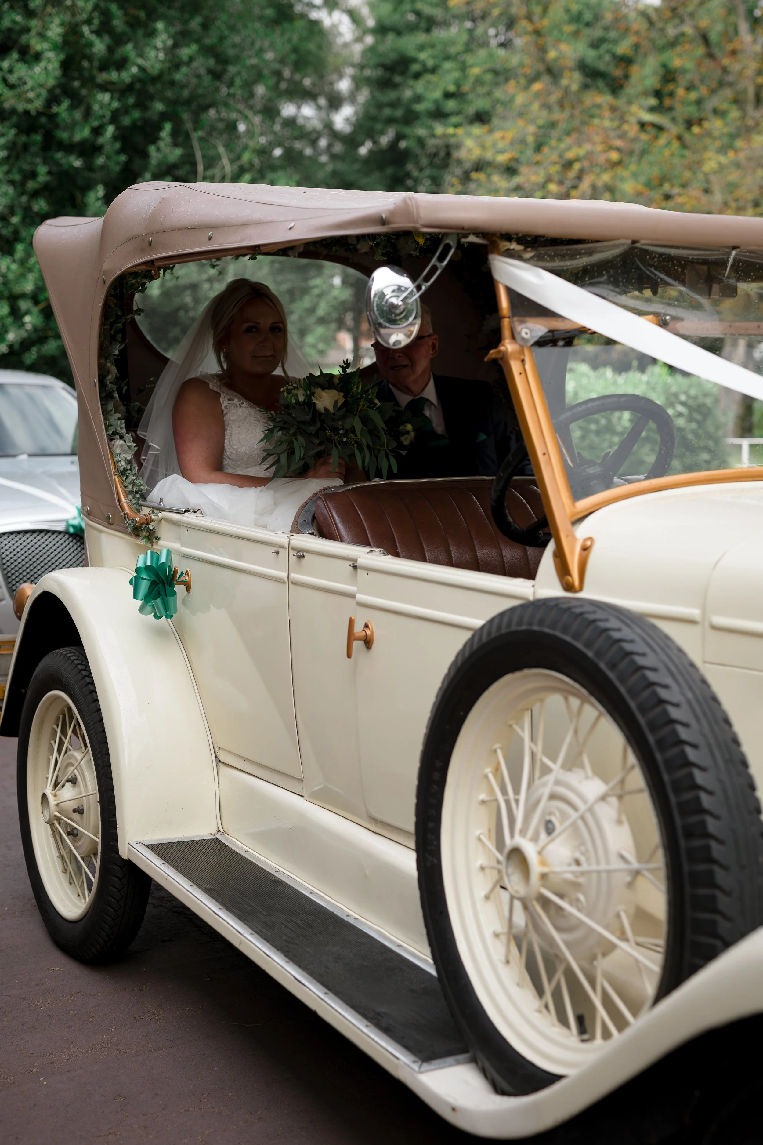 A vintage cream-colored car with a beige convertible top decorated with a green bow, carrying a bride and groom inside, with trees in the background.