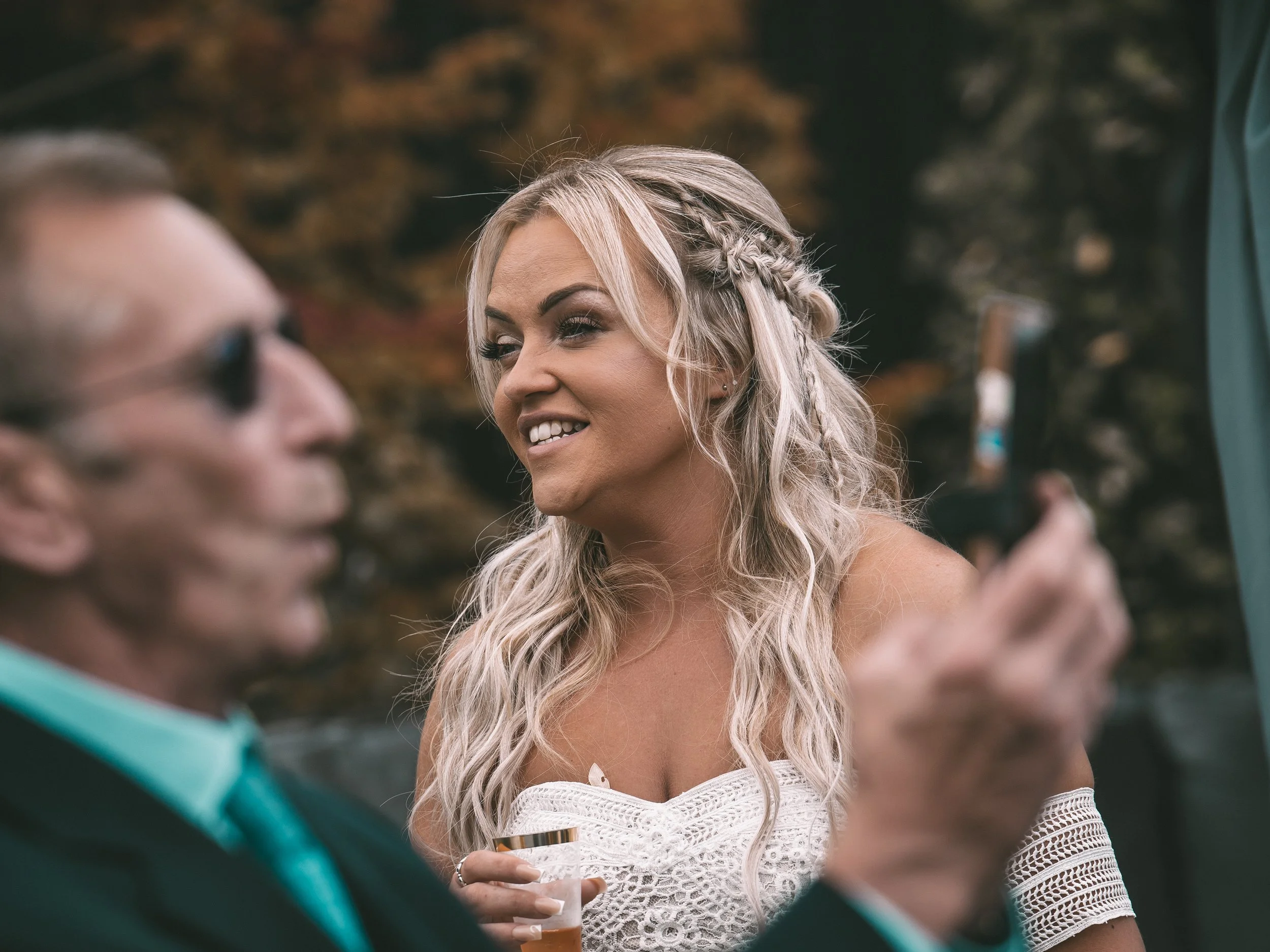 A woman with long, wavy blonde hair and a braided crown smiles while holding a glass of drink. She is wearing a white lace off-shoulder dress. An older man with glasses and a teal shirt is in the foreground, out of focus, engaged in conversation with