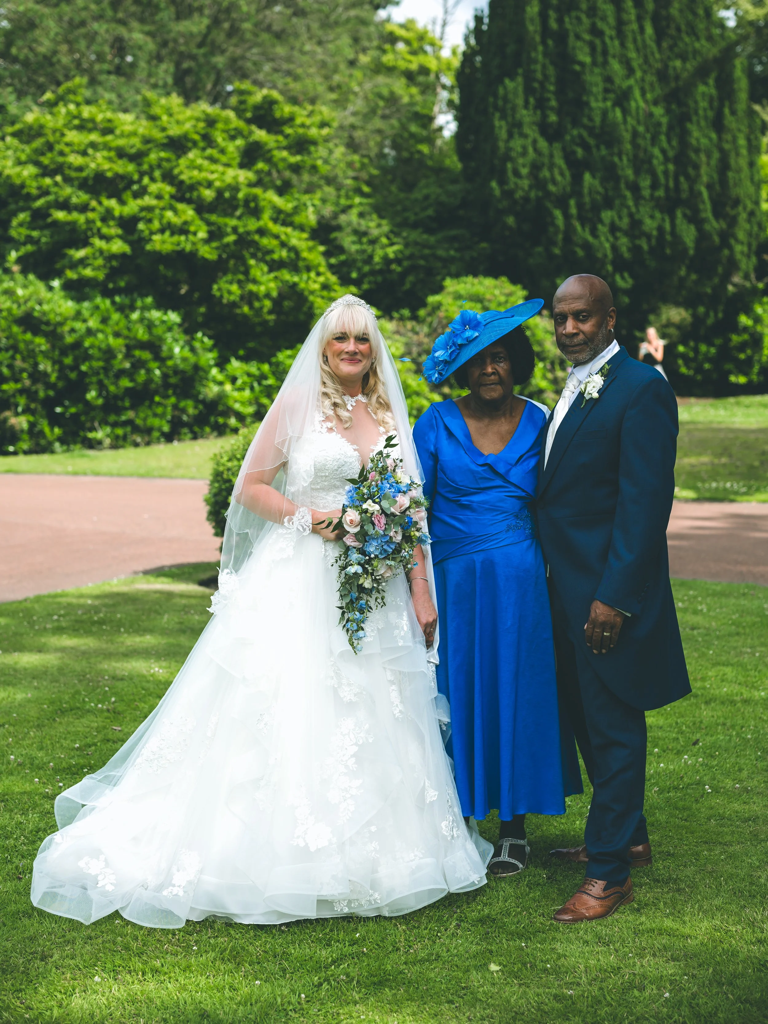 A bride in a white wedding dress with a veil and bouquet, an older woman in a blue dress and matching hat, and a man in a dark suit with boutonniere, standing together outdoors on a grassy lawn with green trees in the background.