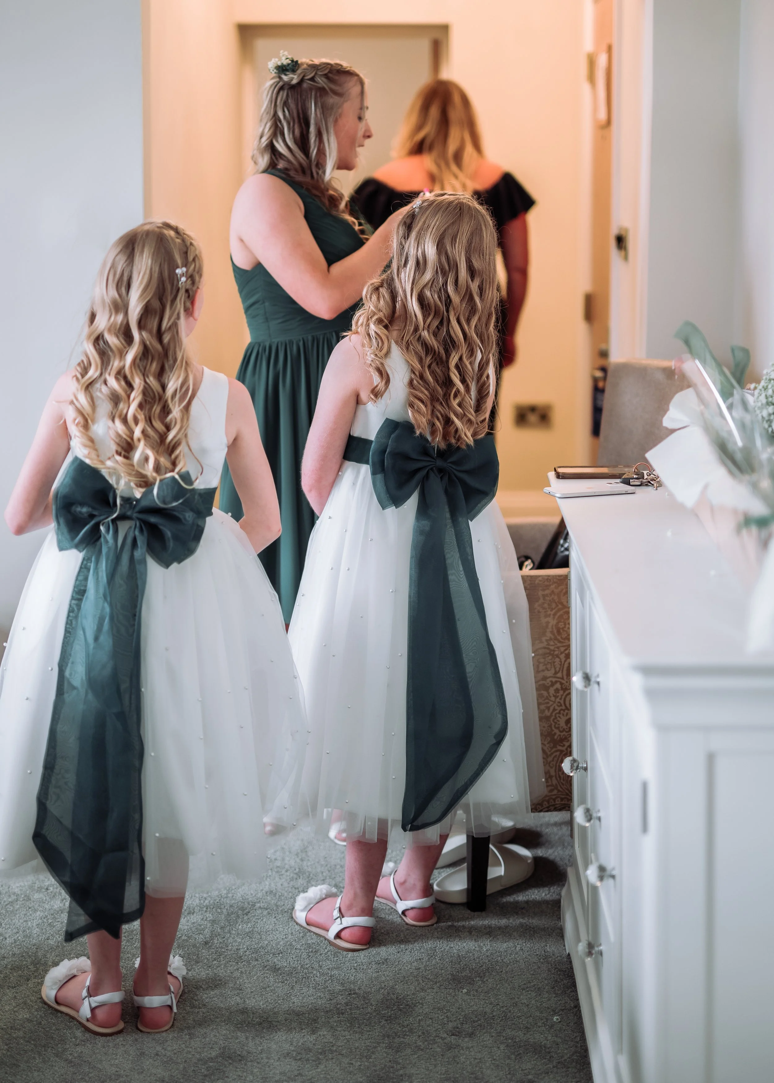 A woman and two young girls with blonde curly hair in white and black dresses with large black bows, standing barefoot and kneeling on a carpet, preparing for a wedding or special event in a cozy room.