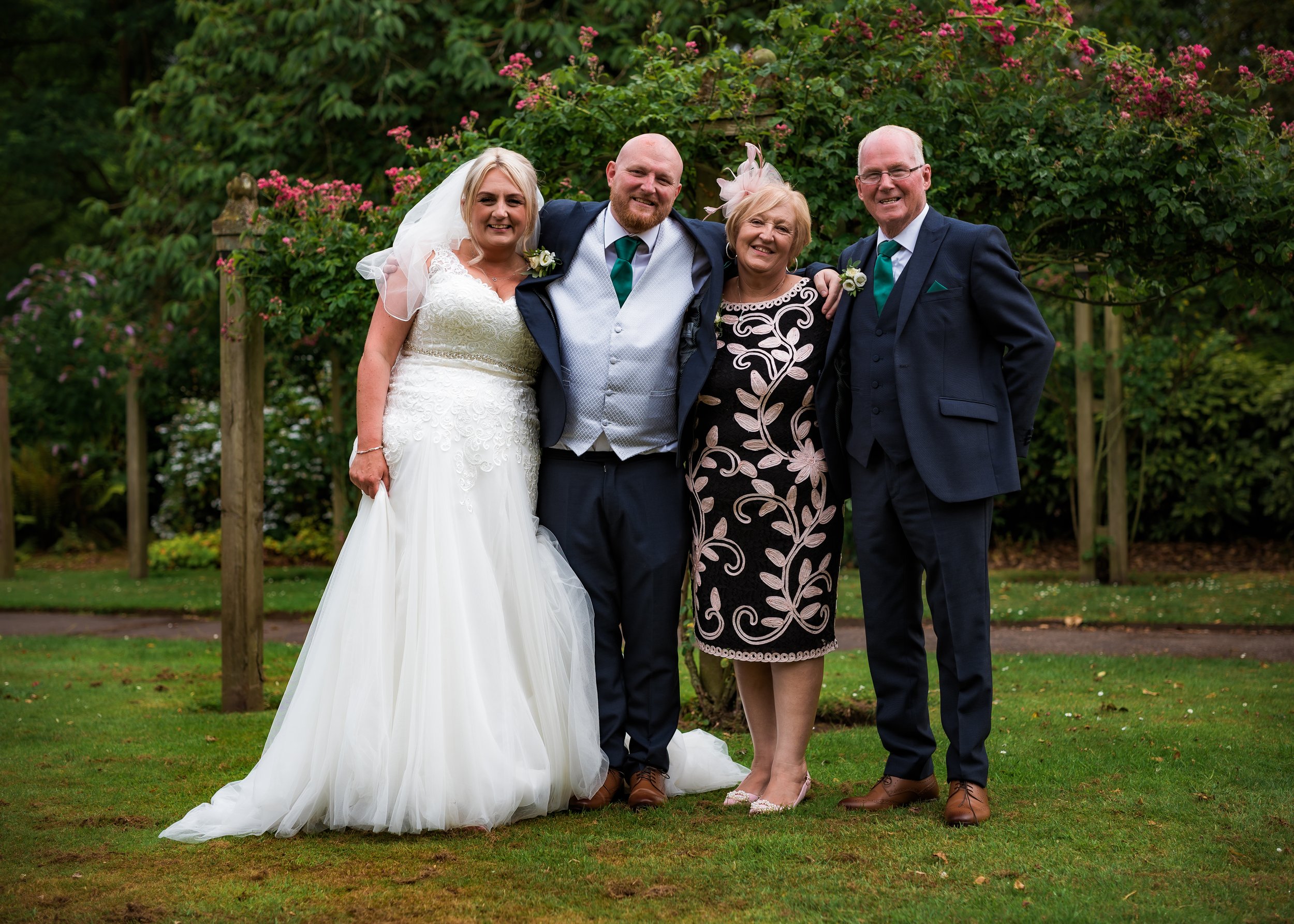 Group of four people at a wedding, standing on grass in front of trees and pink flowers. The bride in a white wedding dress stands next to three older adults in formal suits and dresses, smiling and looking at the camera.