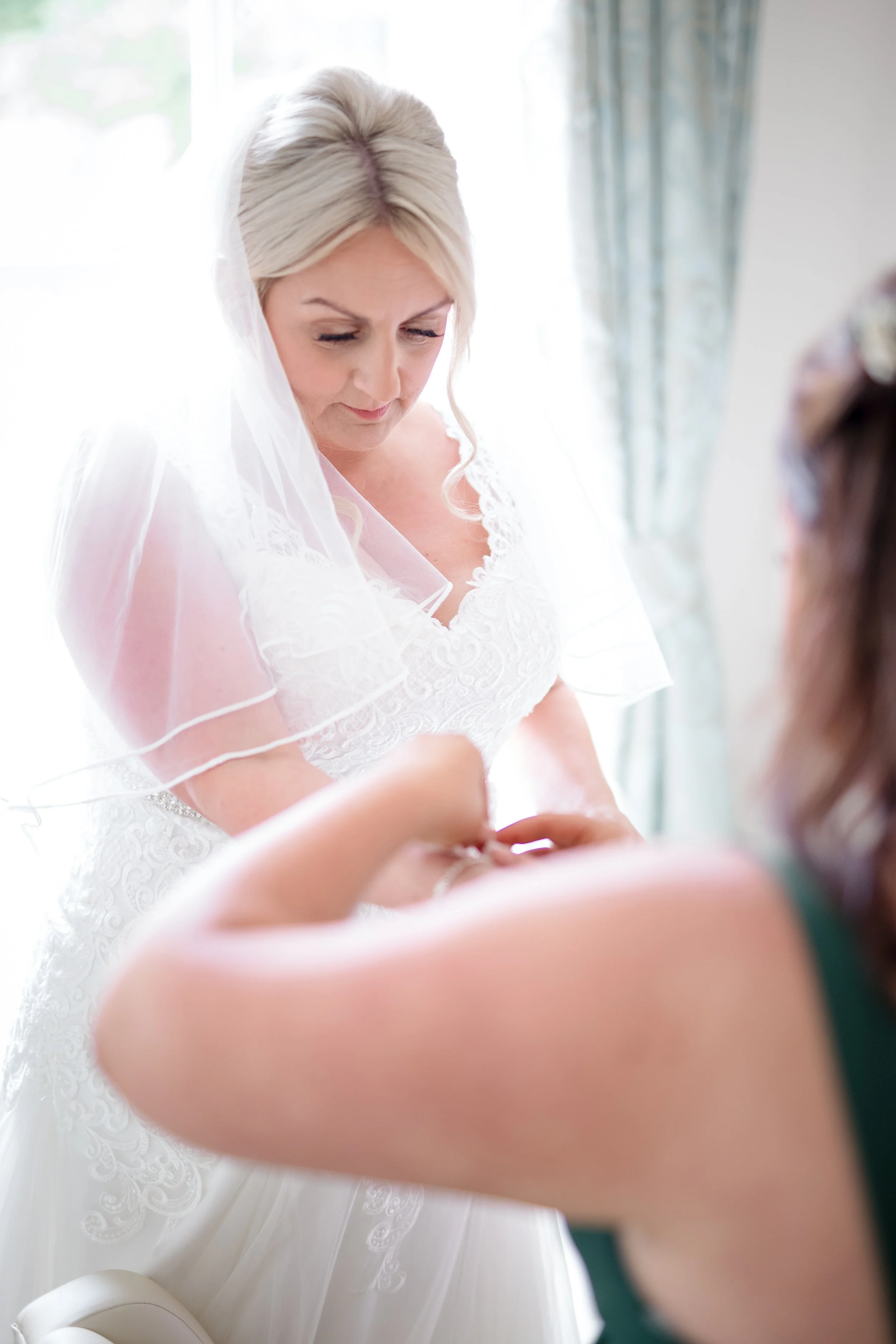 A bride with platinum blonde hair in loose curls wearing a white lace wedding dress and veil, getting ready with assistance from another person in a softly lit room with a window and curtains in the background.