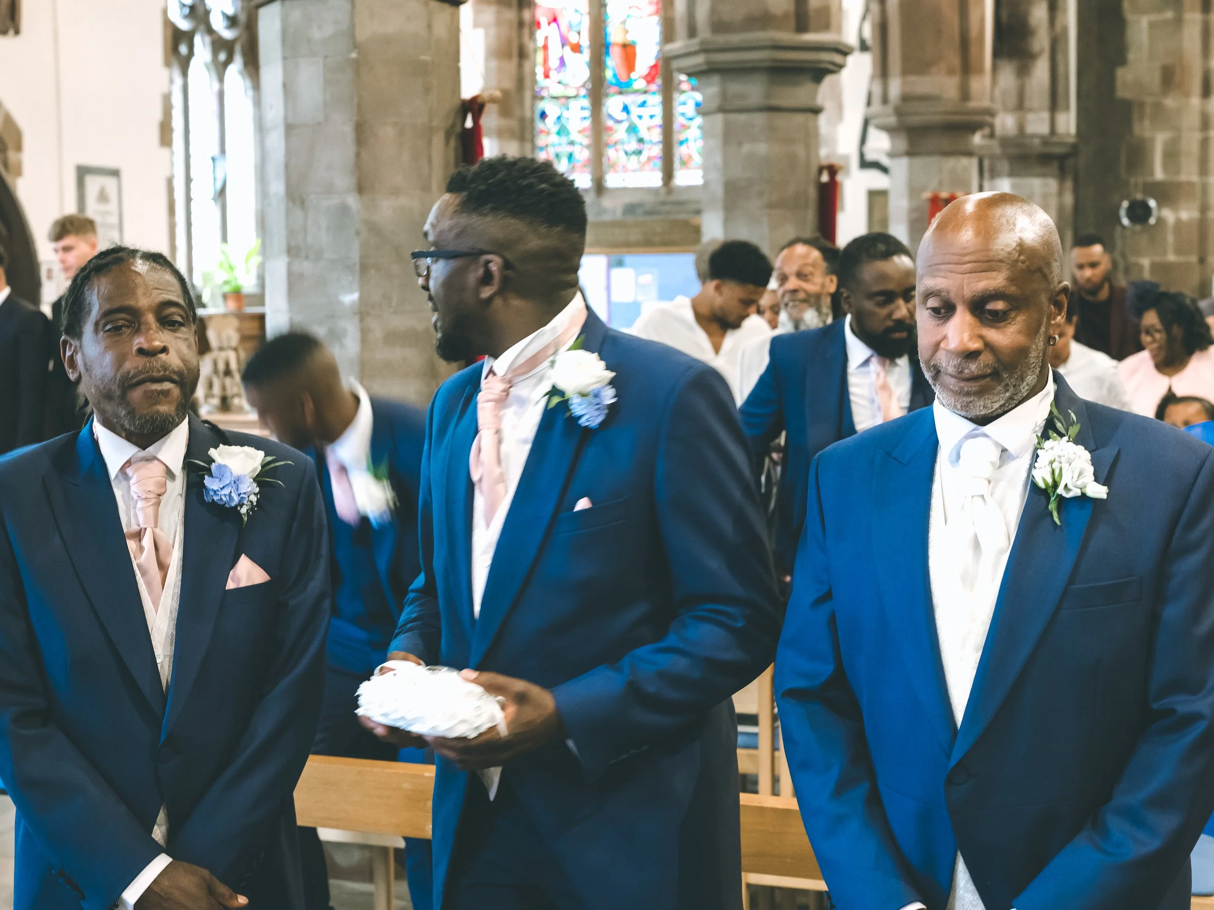 People dressed in formal attire participating in a wedding ceremony inside a church, with stained glass windows in the background.