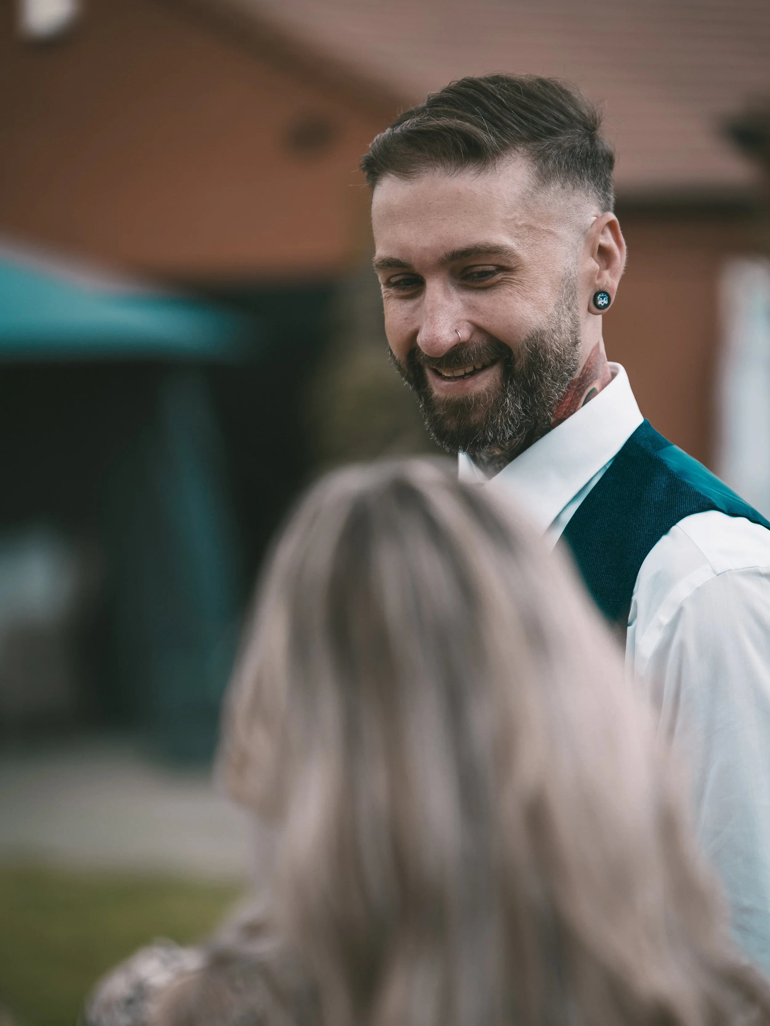 A man with dark hair, a beard, and wearing earrings and a nose ring is smiling and looking down at an outdoor event, with blurred people in the foreground.