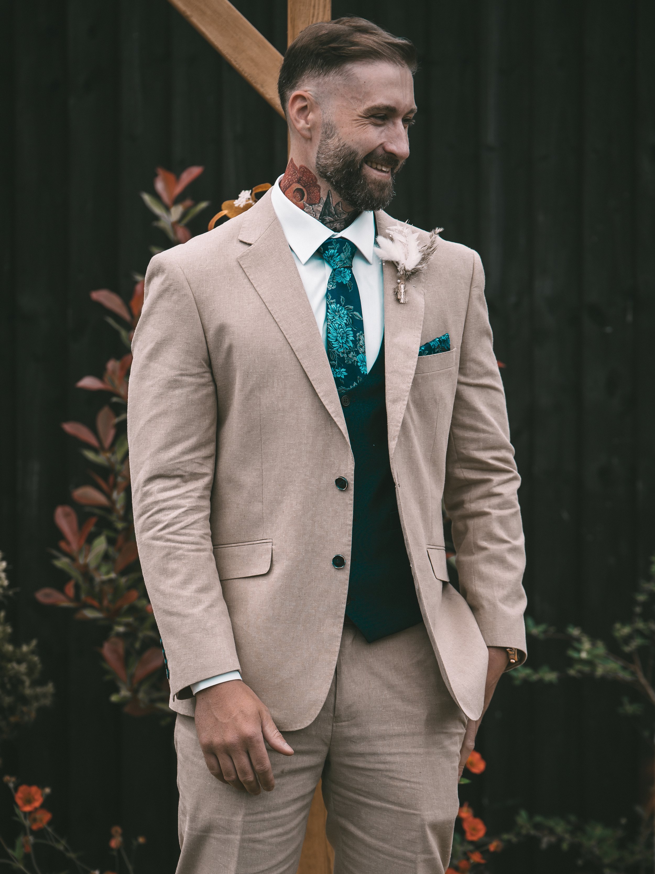 Man wearing a beige suit, white shirt, and blue floral tie, standing outdoors near plants and a black fence, with floral boutonniere on lapel.