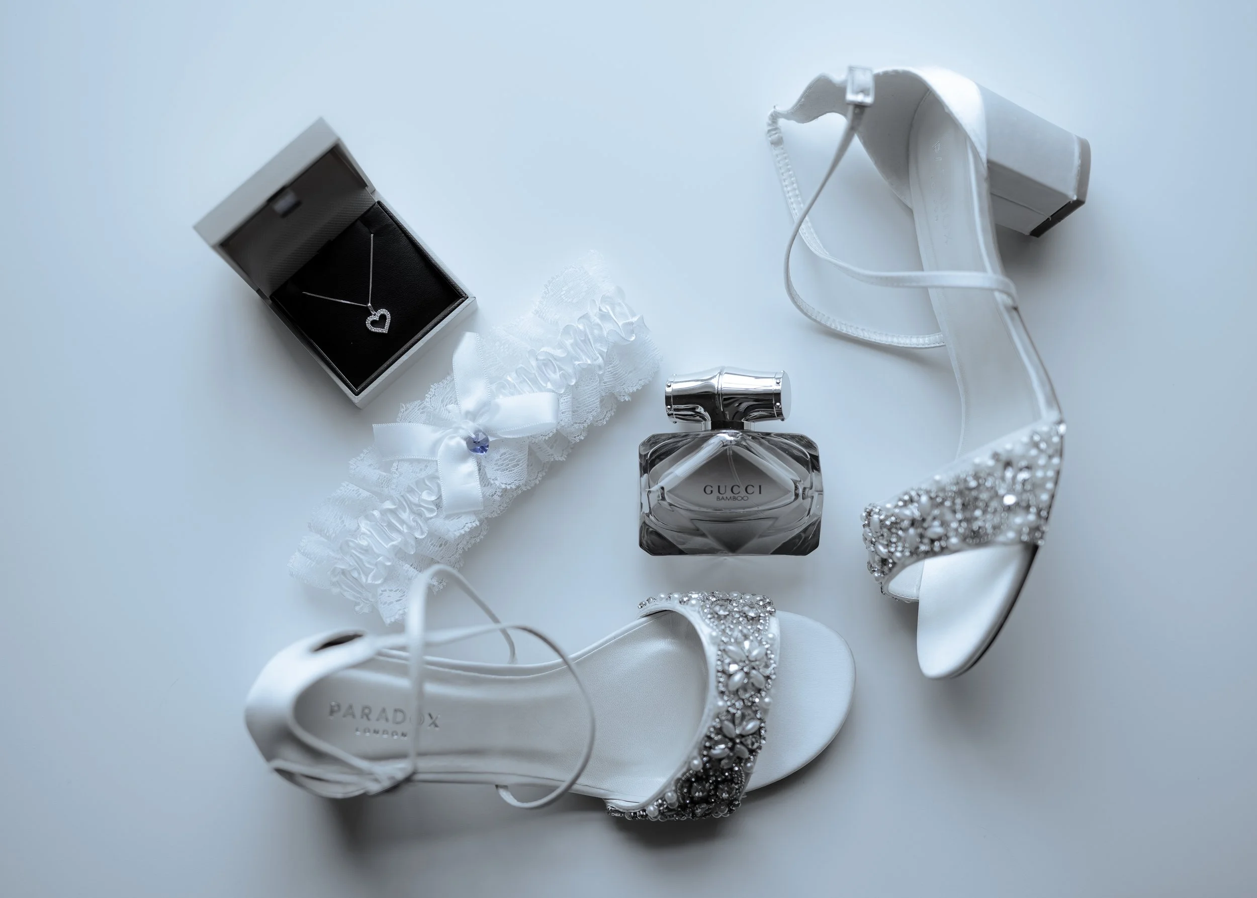 Found jewelry and accessories on a white surface, including a black necklace with a heart pendant inside a box, a white lace hair ribbon, a bottle of Gucci perfume, a white necklace, a pearl scrub bracelet, and a pair of pearl embellished shoes.